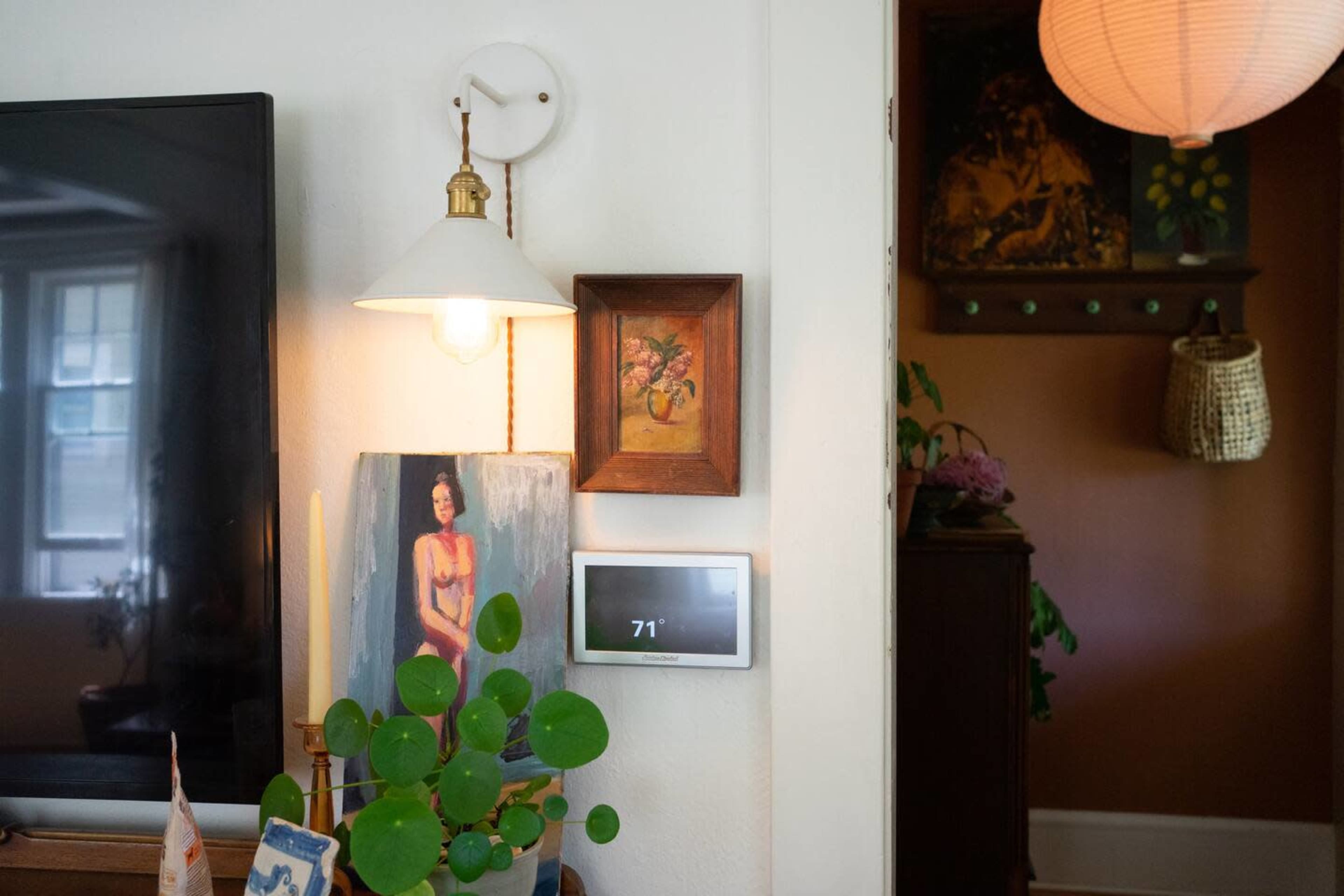 A modern thermostat displays the temperature on a wall next to a painting and a lamp in a cozy room.