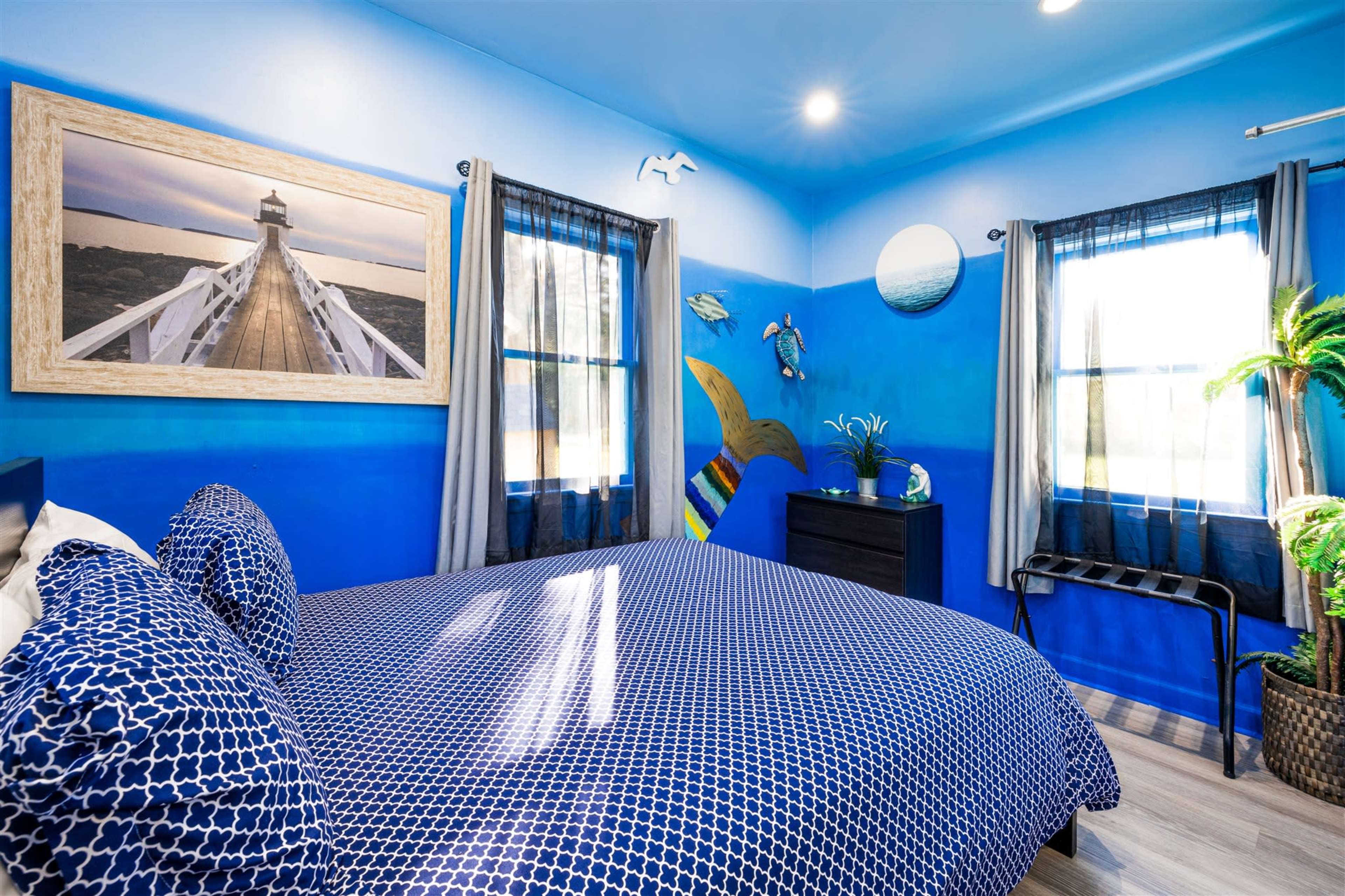 A brightly colored bedroom with blue walls, featuring a double bed with a patterned comforter, a dresser, and artwork of a pier on the wall.