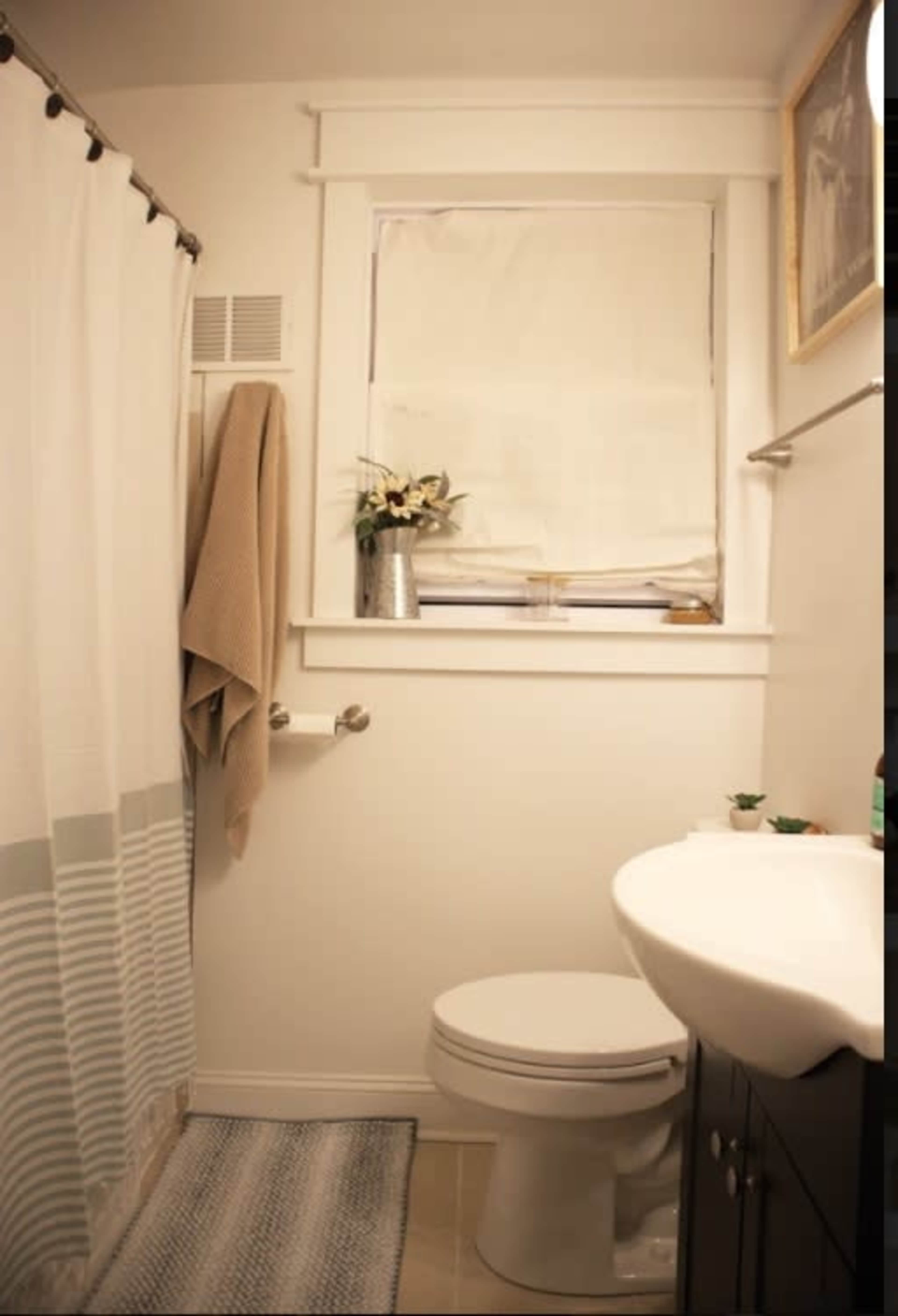 A small bathroom featuring a toilet, sink, window with blinds, a shower curtain, and a decorative towel hung on the wall.