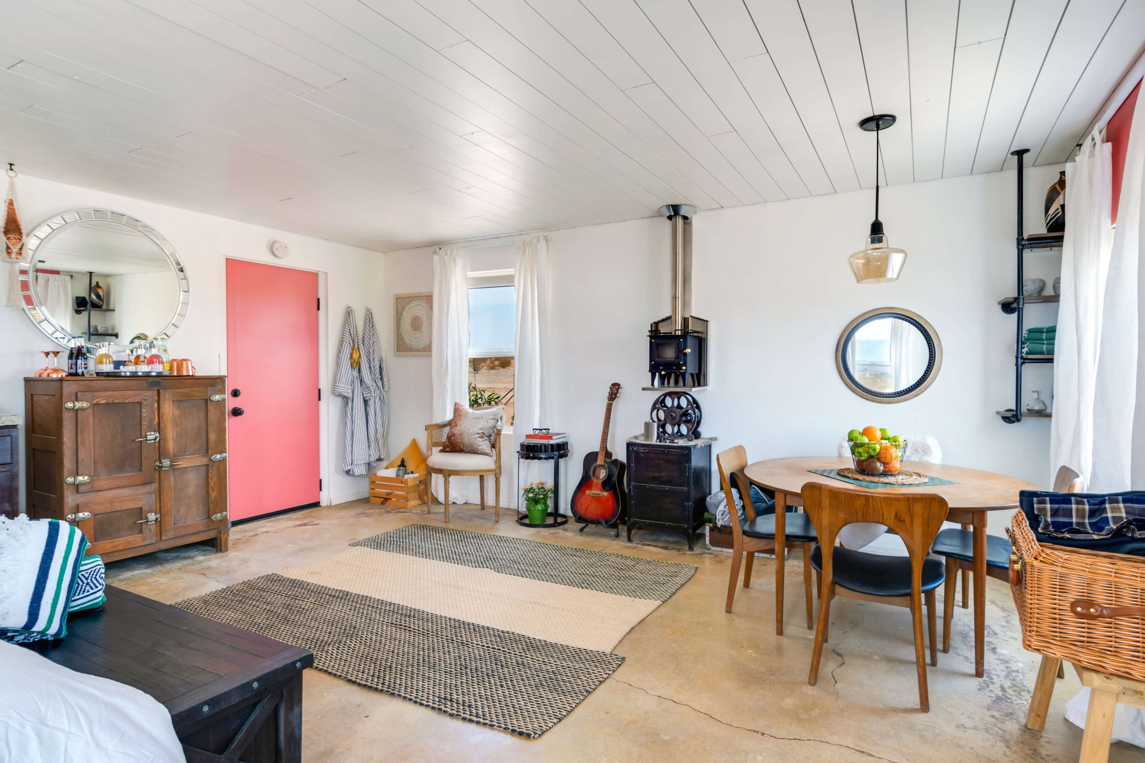 A cozy room with a dining area containing a round table, a guitar, a wood stove, and a pink door, complemented by natural light from large windows.