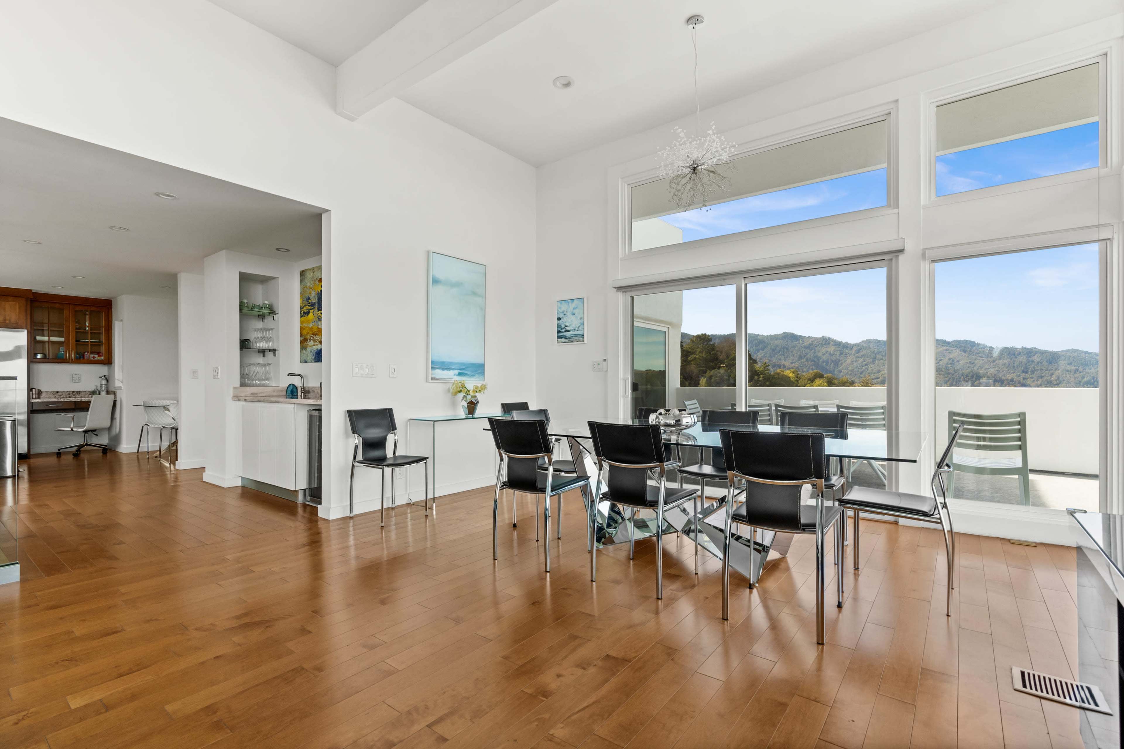 A modern dining area with a glass table and black chairs, adjacent to a kitchen with wooden cabinetry and large windows offering a view of mountains.