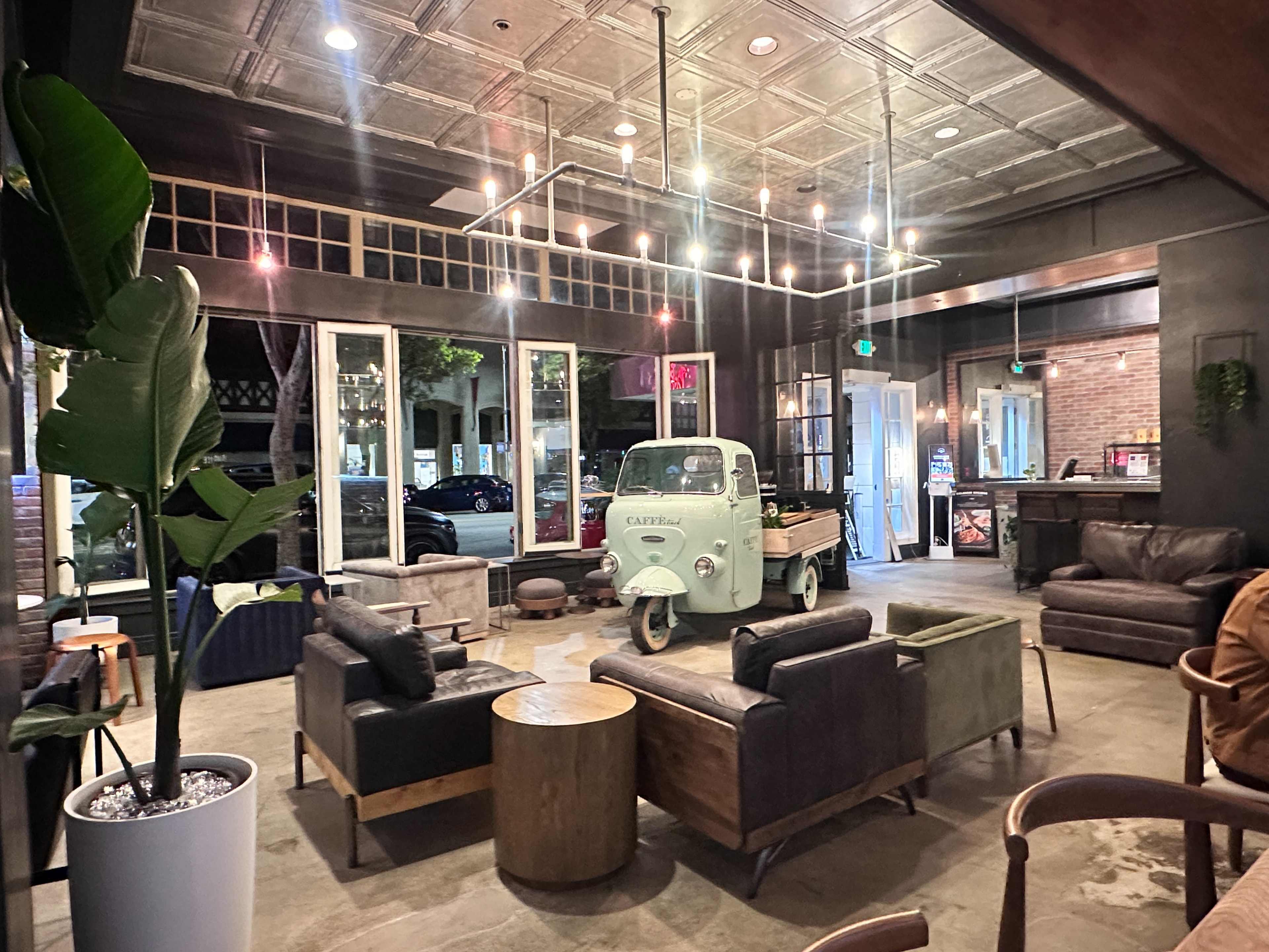 The image shows a modern coffee shop interior featuring a vintage delivery truck as a centerpiece, surrounded by various seating arrangements and large windows.