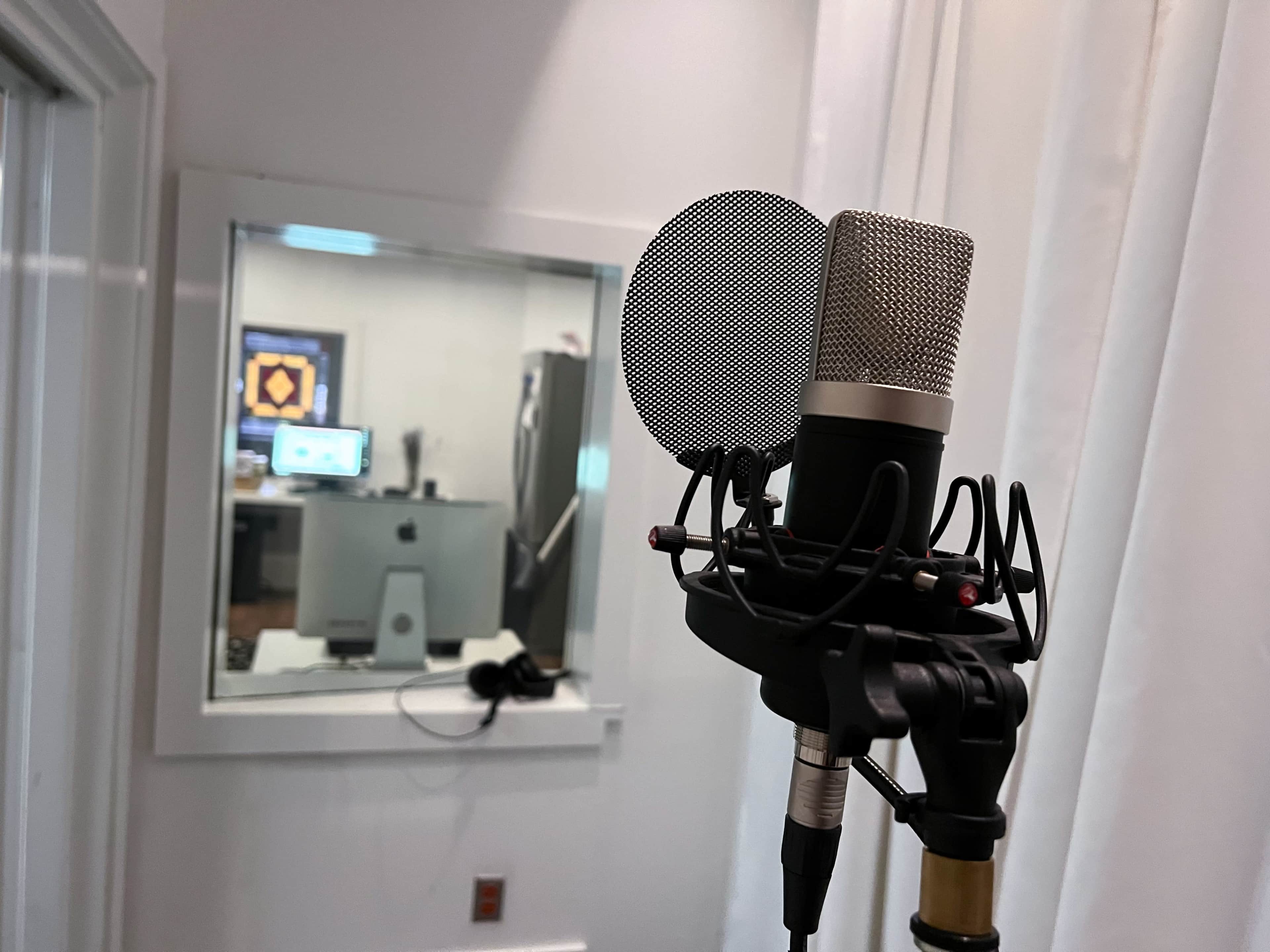 A microphone stands in the foreground while a recording studio is visible through a glass window in the background.