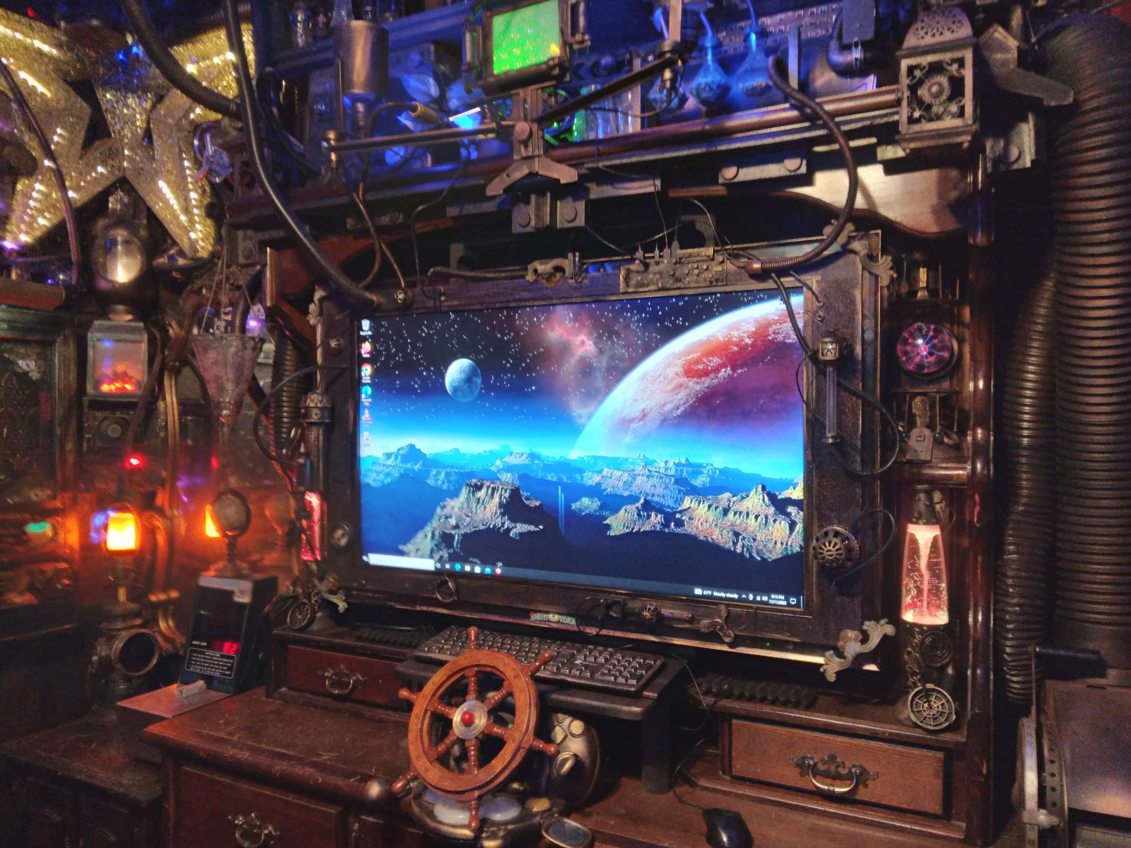 A large computer monitor displaying a space-themed wallpaper is surrounded by an intricate setup of steampunk-inspired decorations and devices.