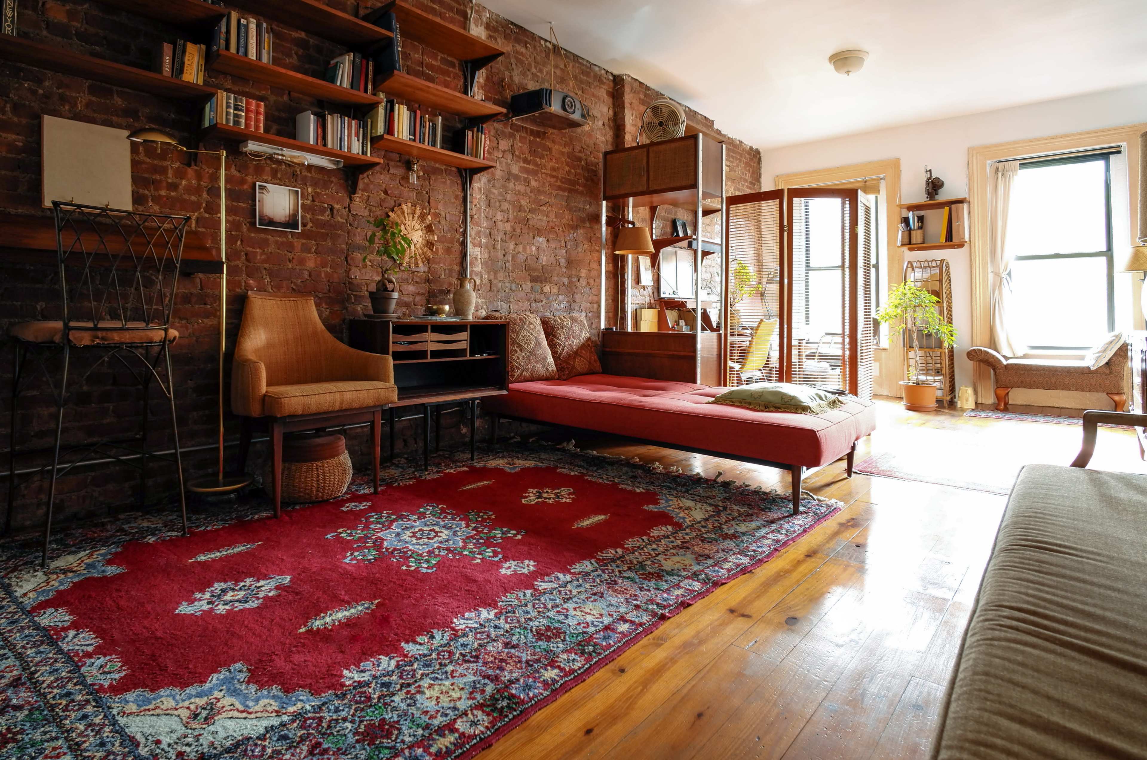 The image shows a cozy living room with exposed brick walls, wooden floors, a red area rug, and various plants and furniture arranged in a minimalist style.