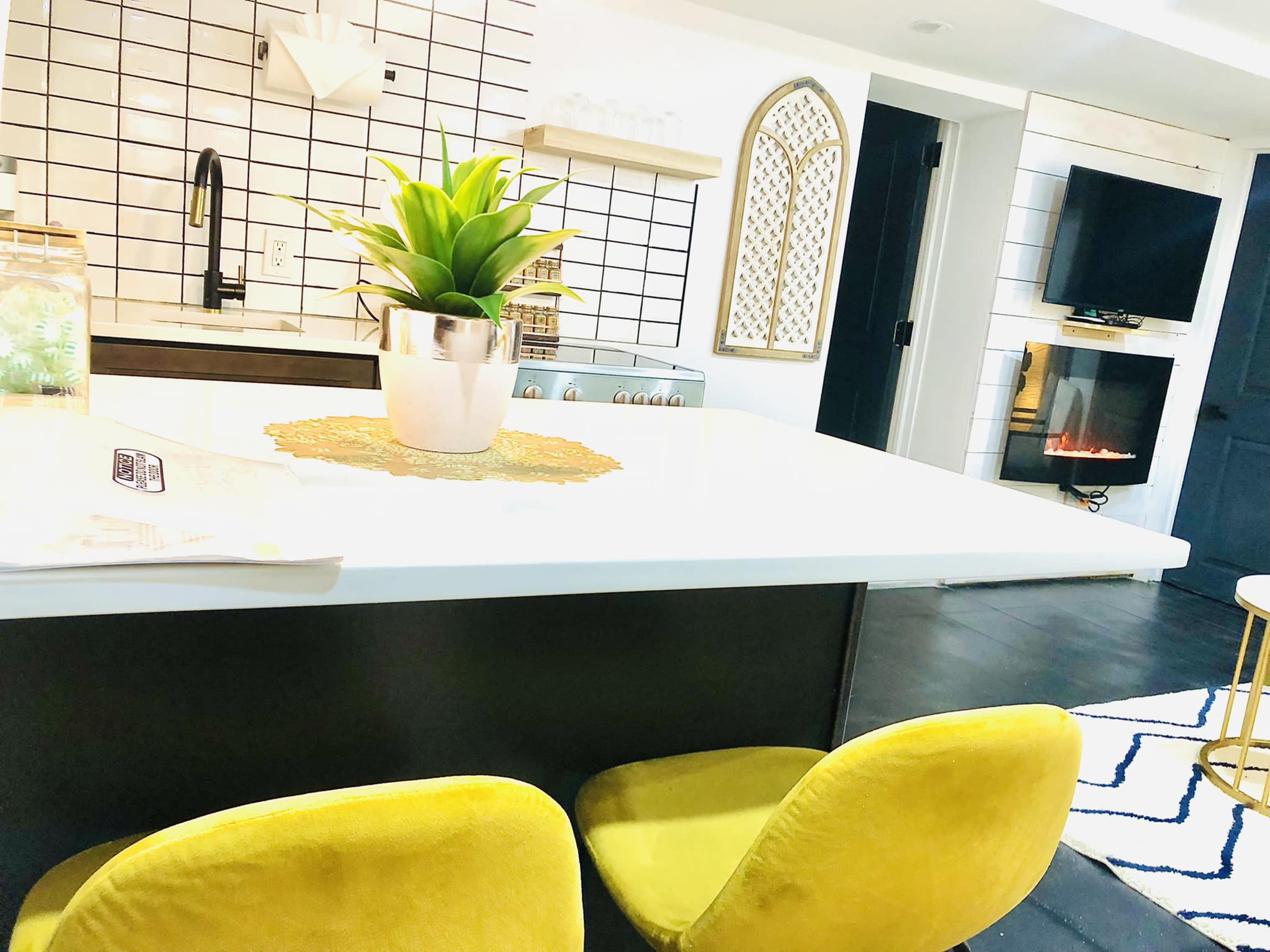 The image shows a modern kitchen with a white countertop, yellow upholstered chairs, a potted plant, and a wall-mounted television.