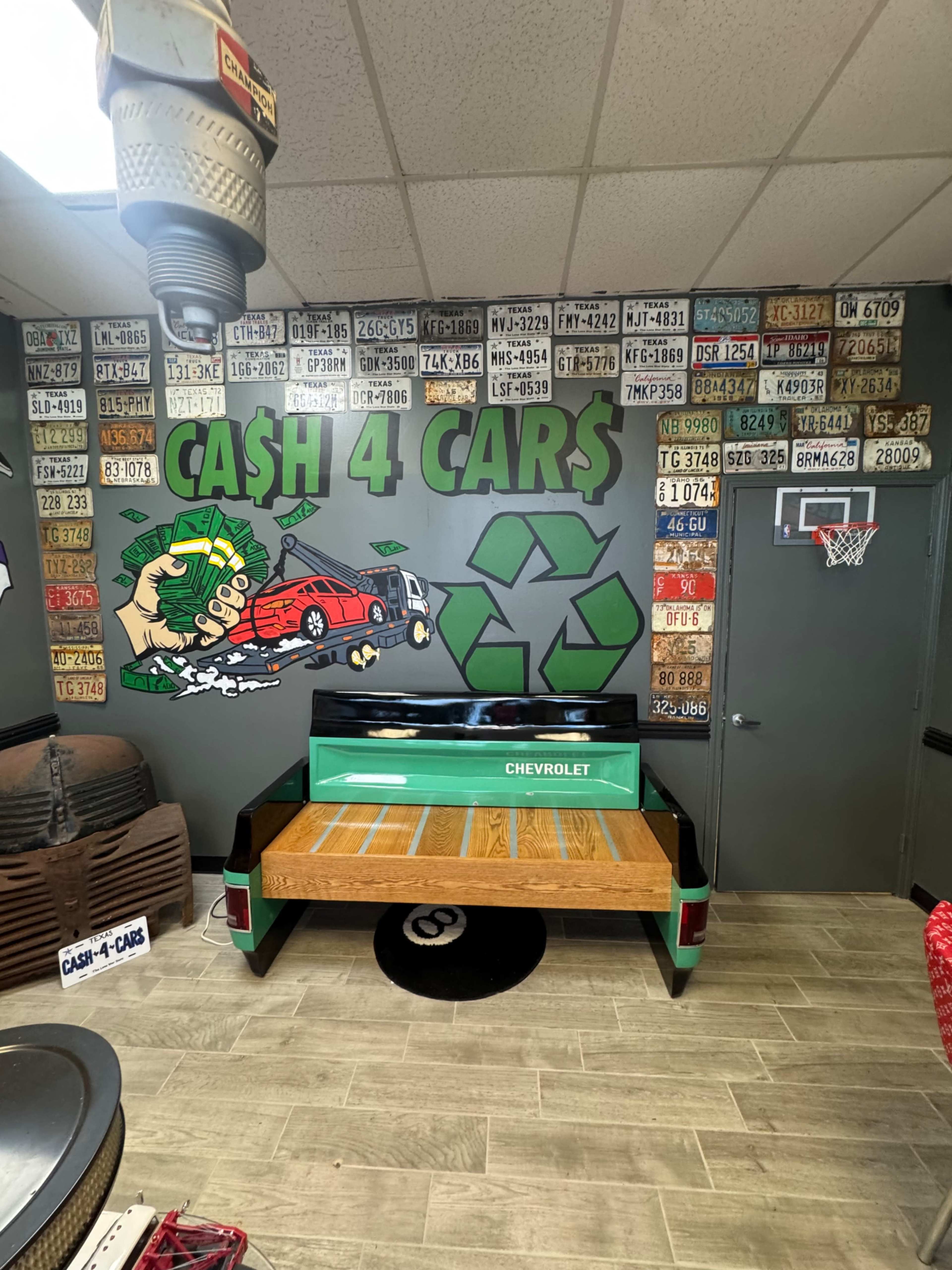 The image shows a room with a mural featuring a car and a recycling symbol on the wall, surrounded by various license plates, and a green and wood platform resembling a Chevrolet truck bed.