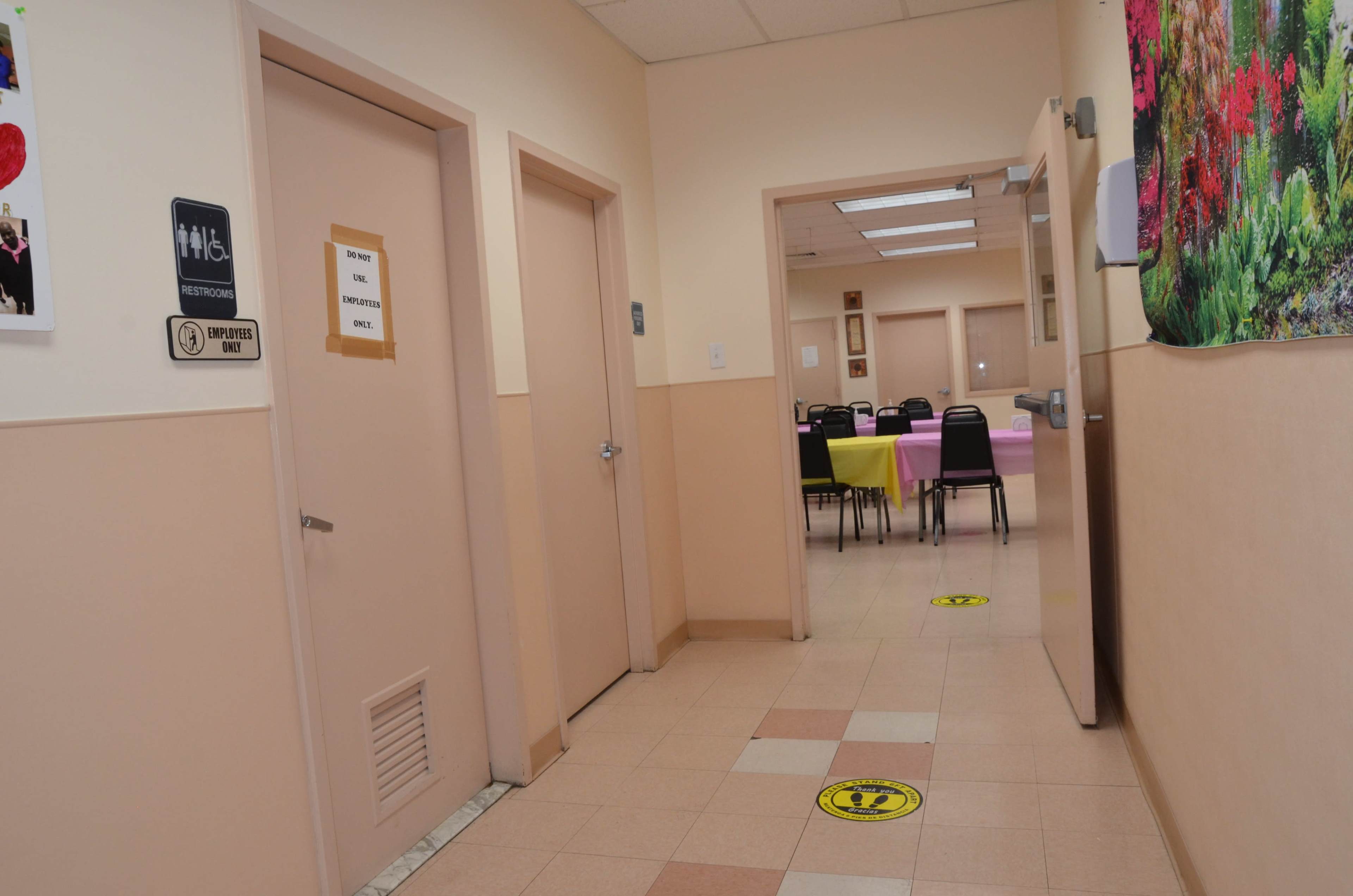 A hallway with two doors on the left and a dining area visible at the end, featuring tables covered with pink and yellow tablecloths.