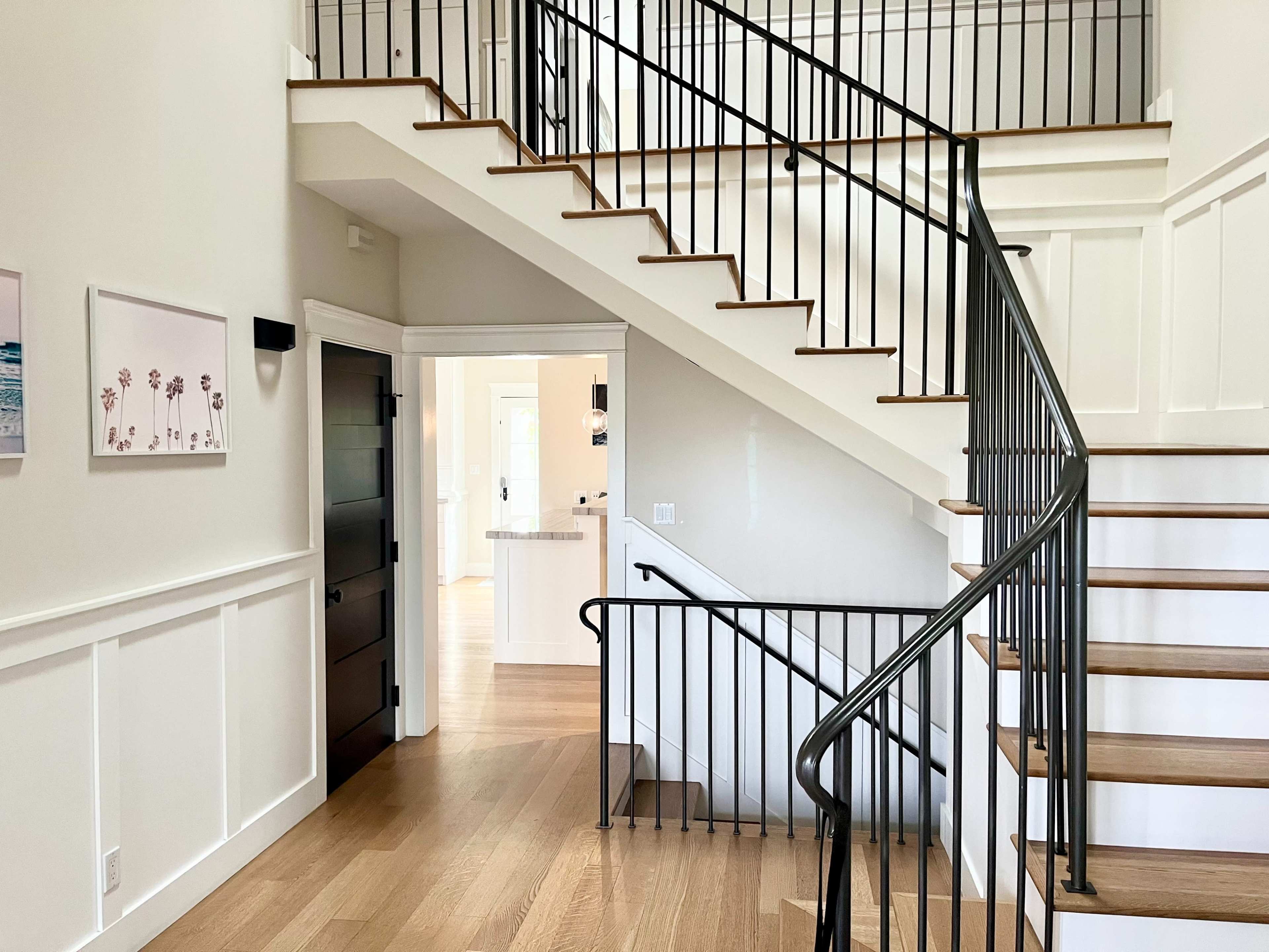 The image depicts a well-lit entryway featuring a spiraling staircase with wooden steps and black metal railings, surrounded by light-colored walls and minimal decor.