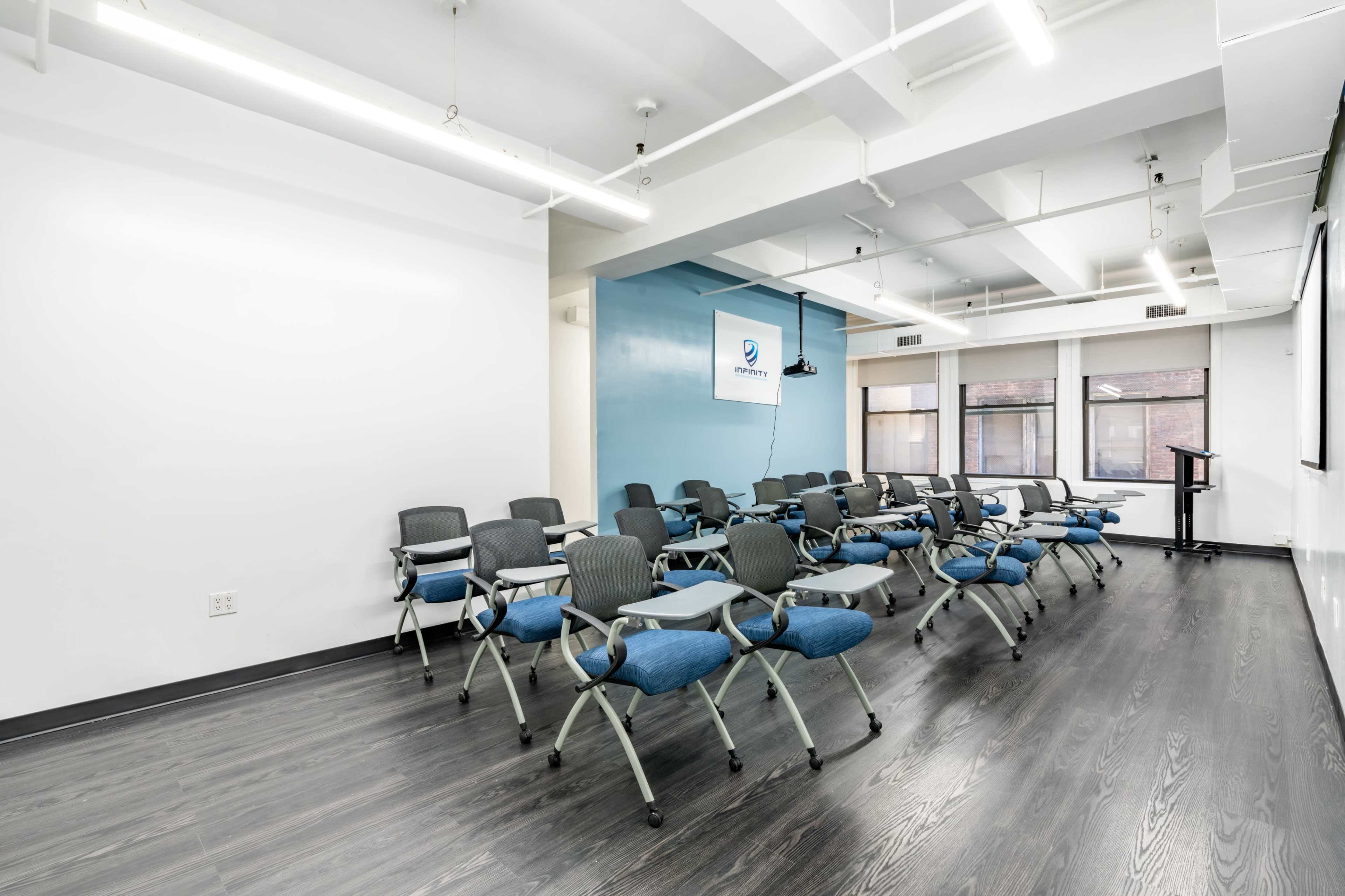 A conference room features rows of blue and black chairs set up for a meeting, with large windows and a presentation screen at the front.
