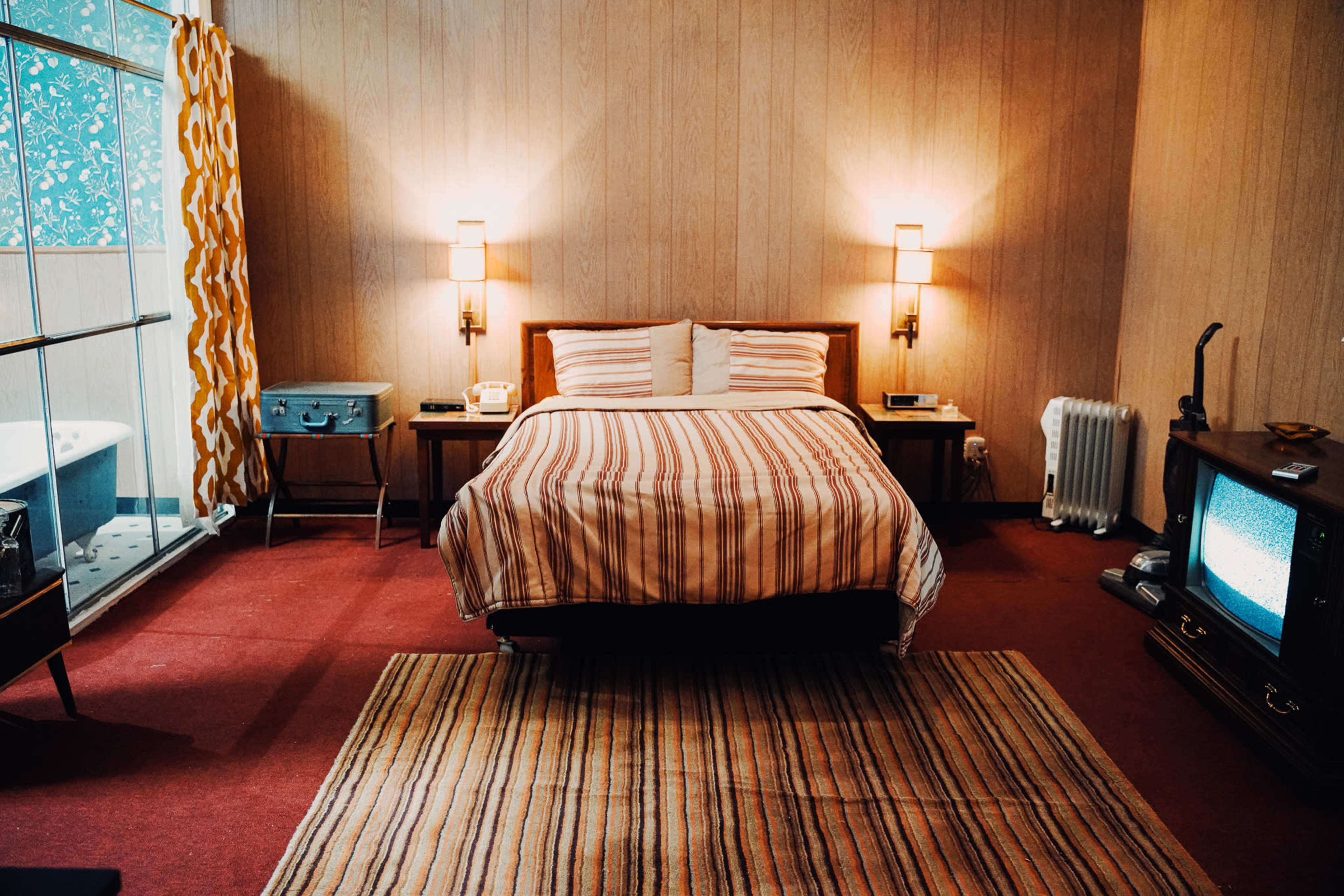 A retro-style hotel room featuring a bed with striped bedding, a patterned carpet, and a vintage television placed next to a wooden nightstand.