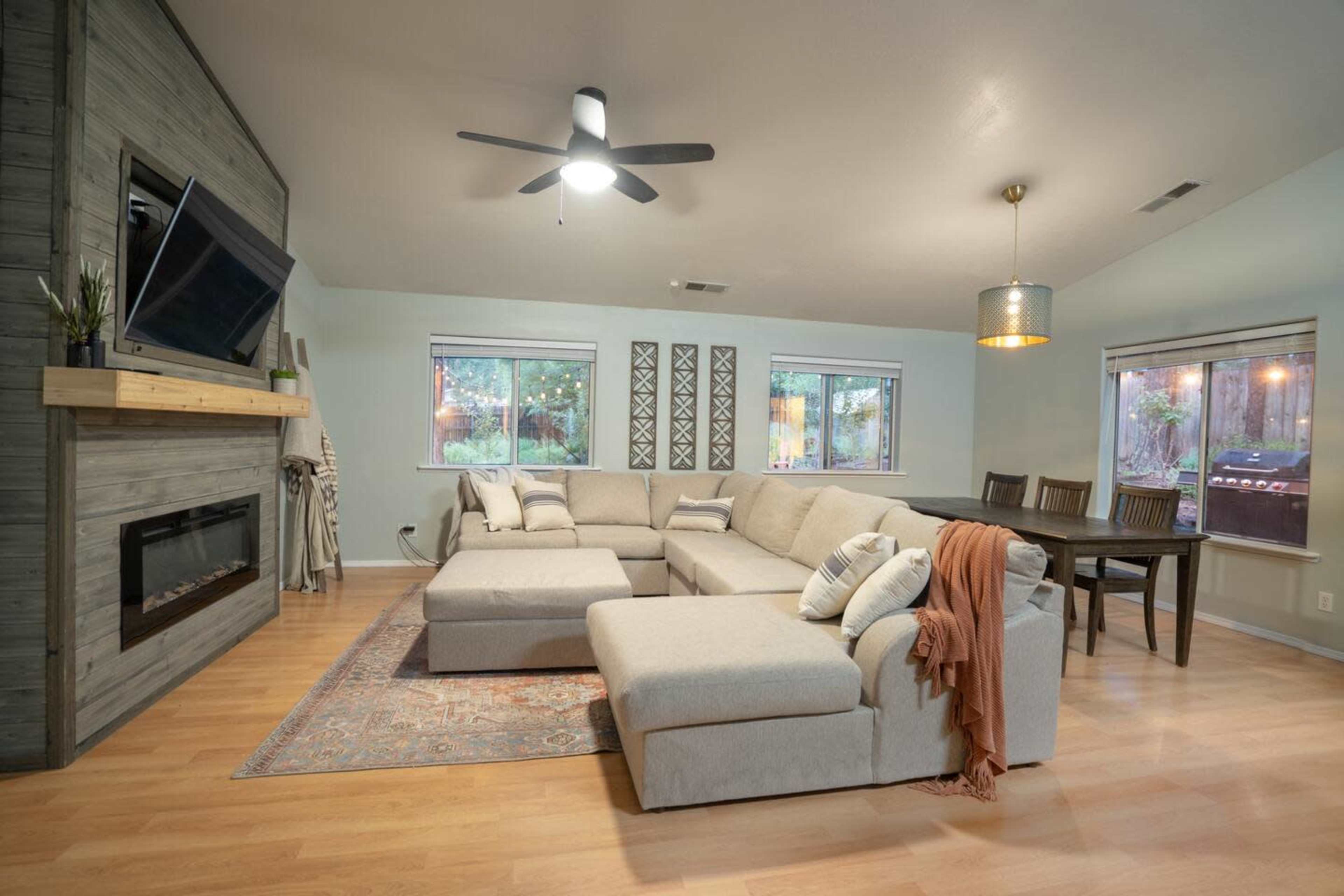 The image shows a spacious living room with a large sectional couch, a wall-mounted television, a fireplace, and a dining area with a wooden table and chairs.
