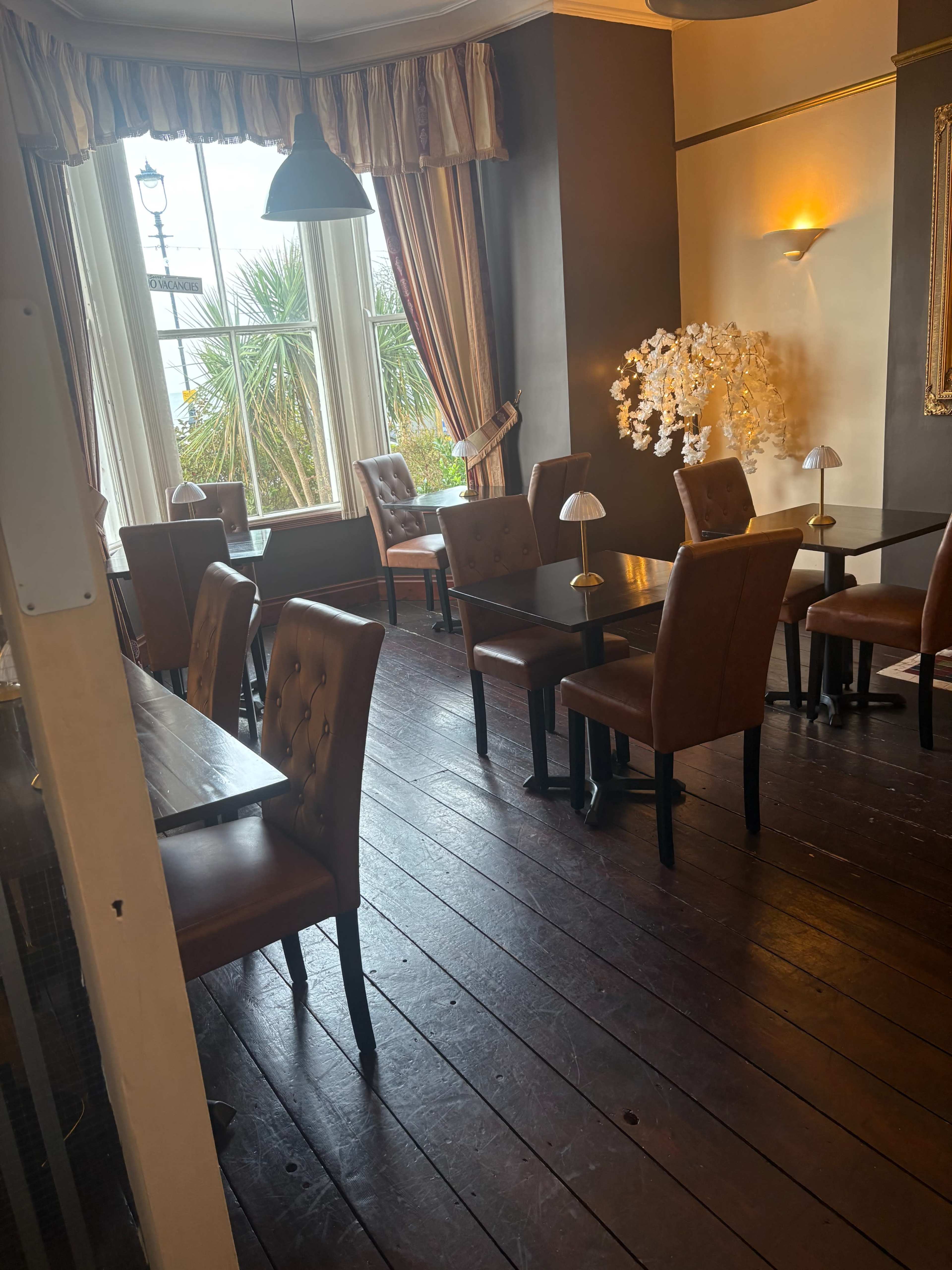 The image shows a cozy dining area with brown leather chairs and wooden tables, illuminated by soft lighting and featuring large windows.
