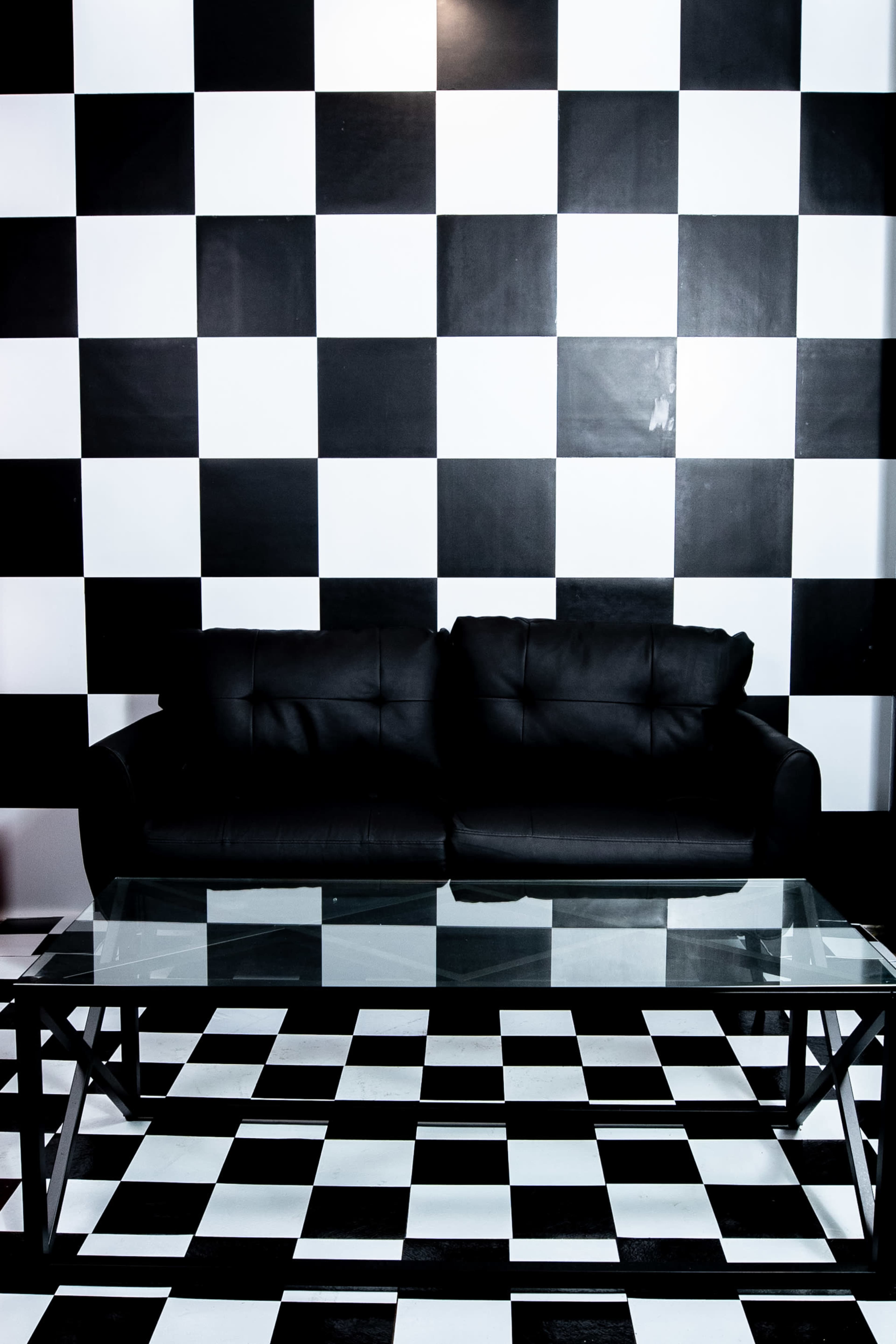 A black leather sofa sits in front of a glass coffee table on a checkered black and white floor, with a matching checkered wall behind it.