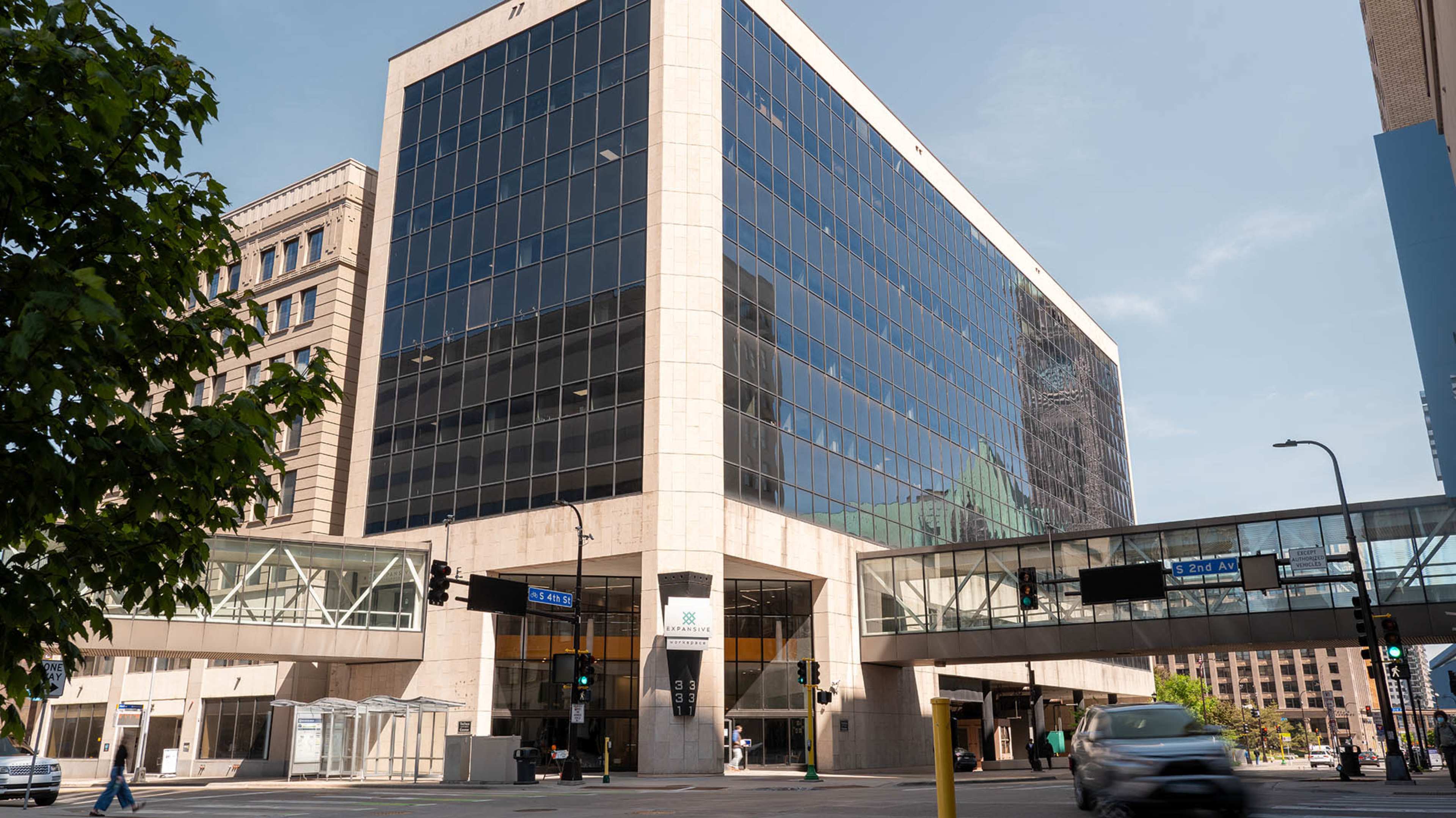 The image shows a modern glass-fronted building at an urban intersection, with a pedestrian bridge connecting to another structure.