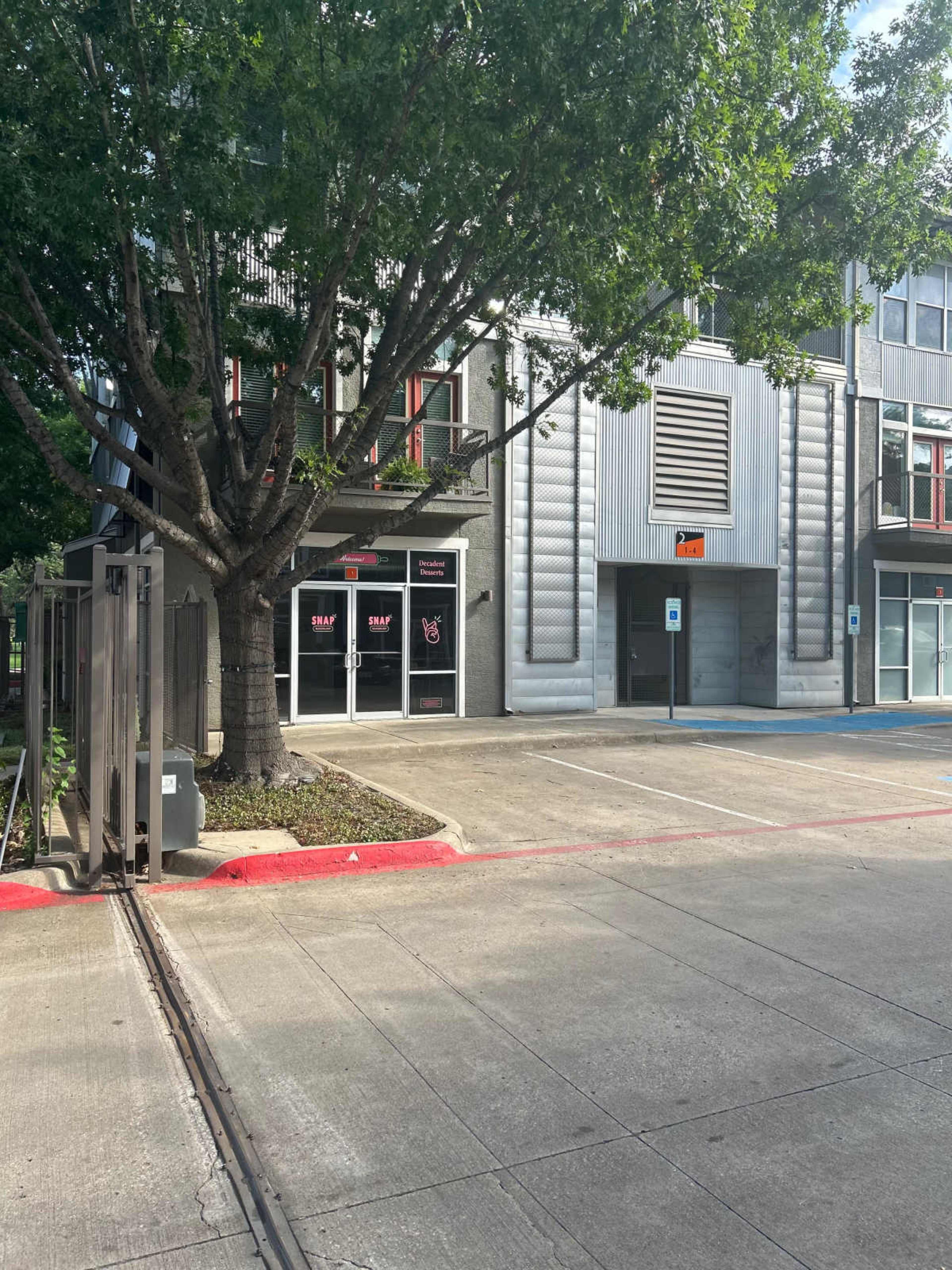 The image shows a parking area in front of a multi-story building with large windows and a prominent tree beside it.