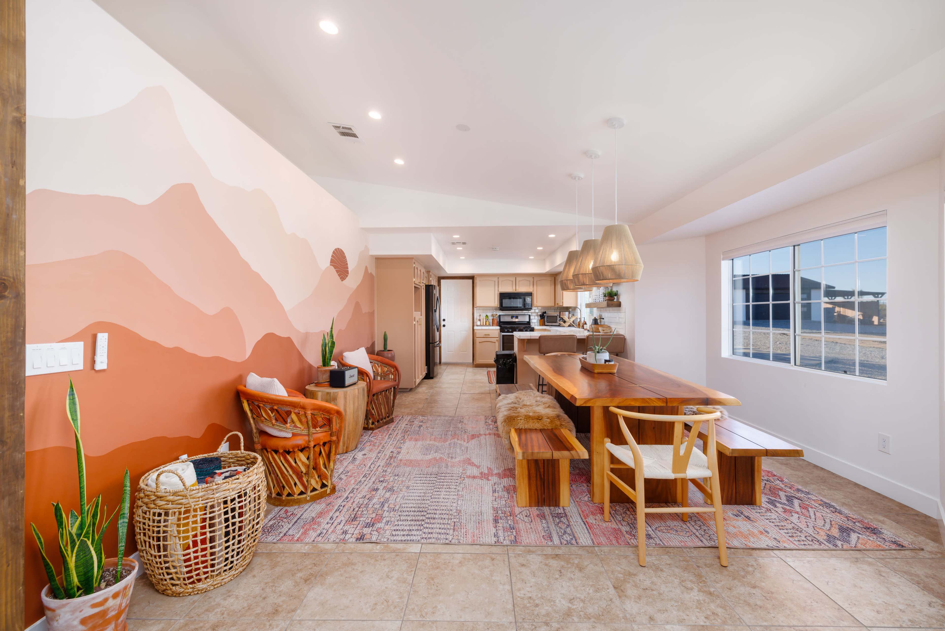 The image shows a modern dining area with a long wooden table, surrounded by chairs and featuring a large wall mural with abstract mountain shapes in warm colors.