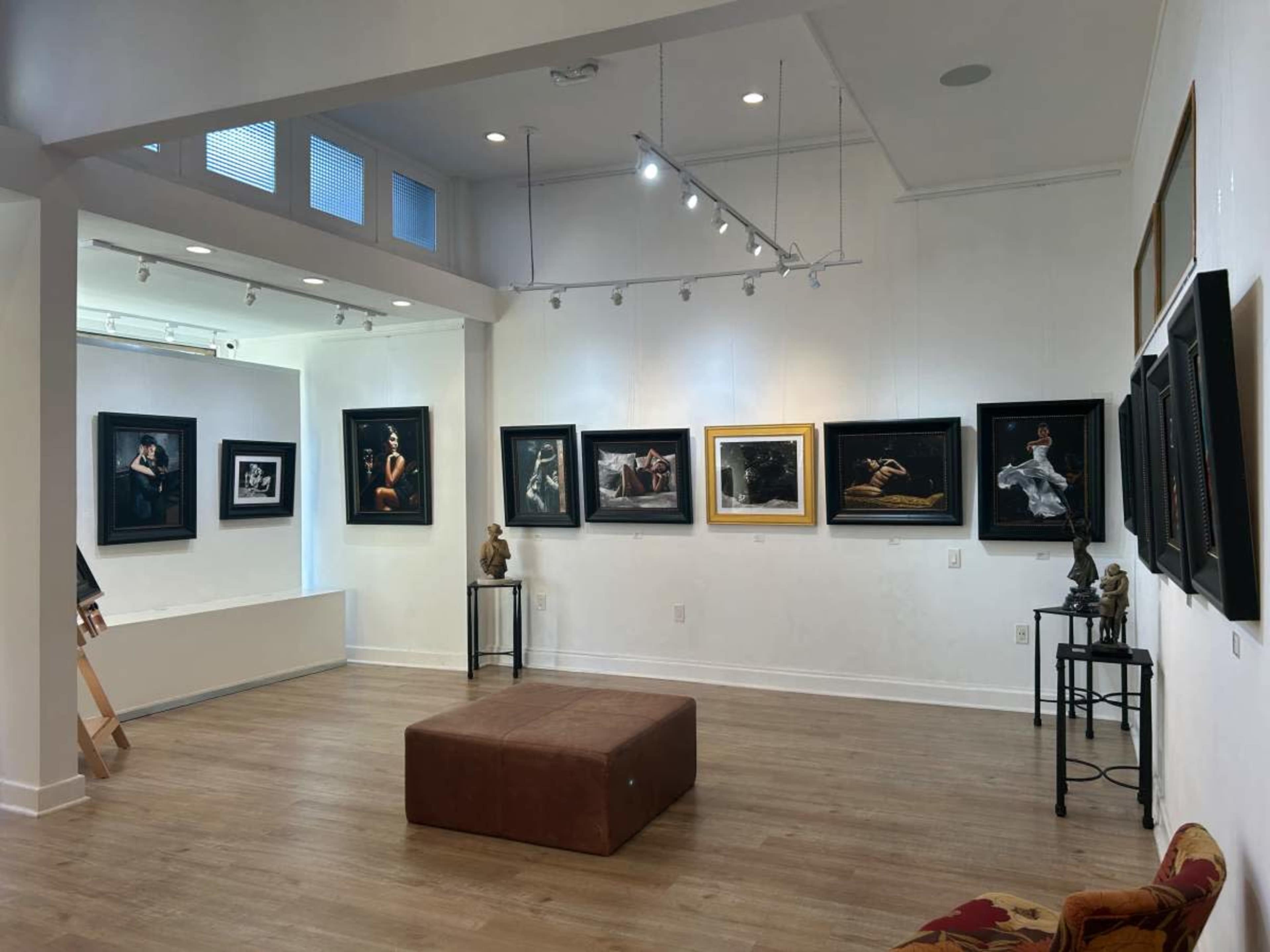 The image depicts a well-lit art gallery showcasing framed photographs and sculptures displayed against white walls and wooden flooring.