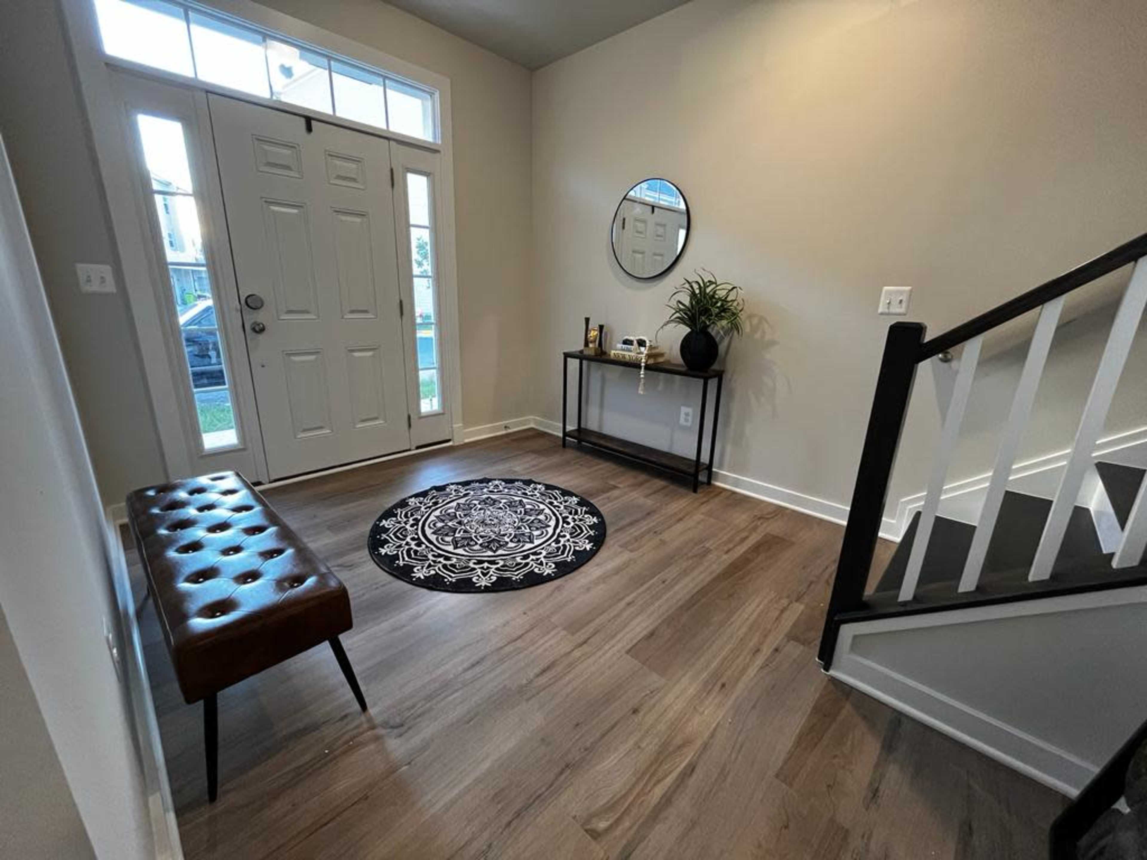 A modern entryway featuring a leather bench, a round rug, a circular mirror, and a small table against a wall, with a staircase visible in the background.