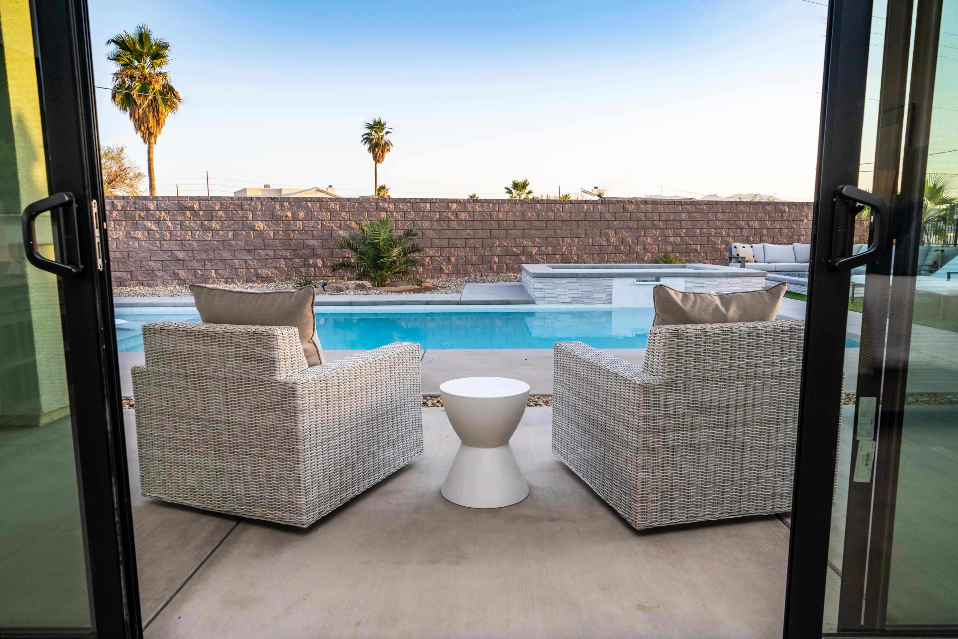 A pair of white wicker chairs and a small round table face a swimming pool surrounded by palm trees and a stone wall.