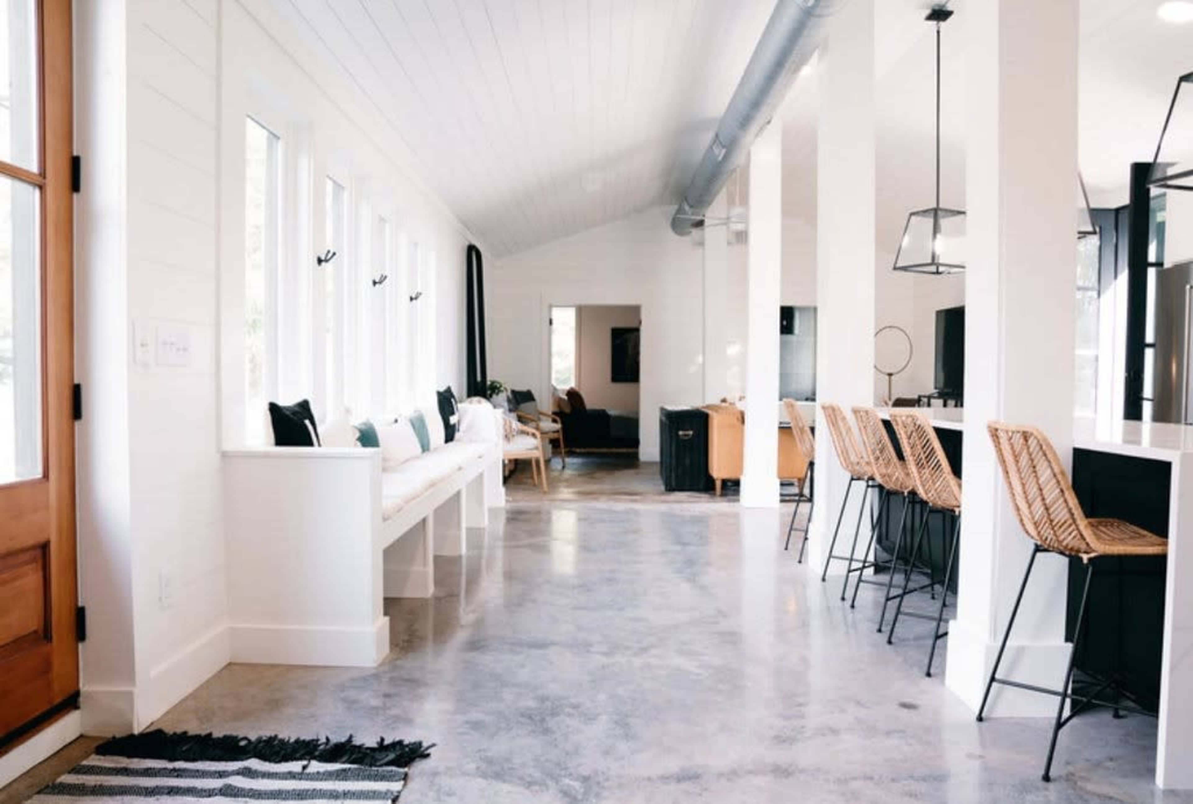 The image shows a modern, open-concept living space with a polished concrete floor, large windows, and a kitchen counter with bar stools.