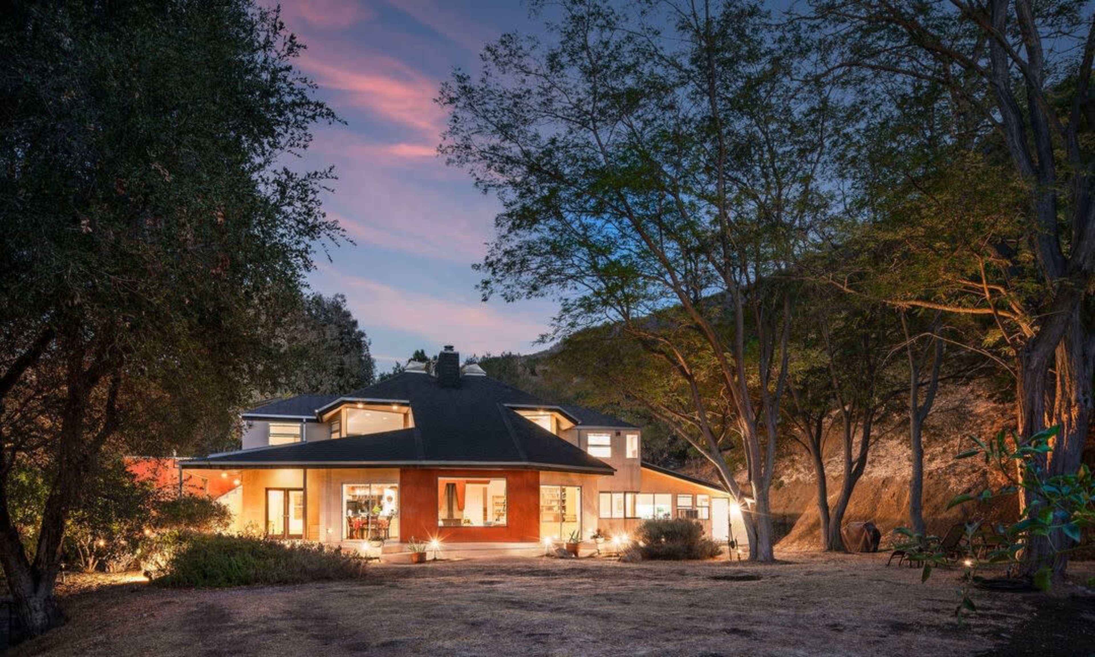 A modern house with large windows is set amid trees at dusk, illuminated by soft interior lights.