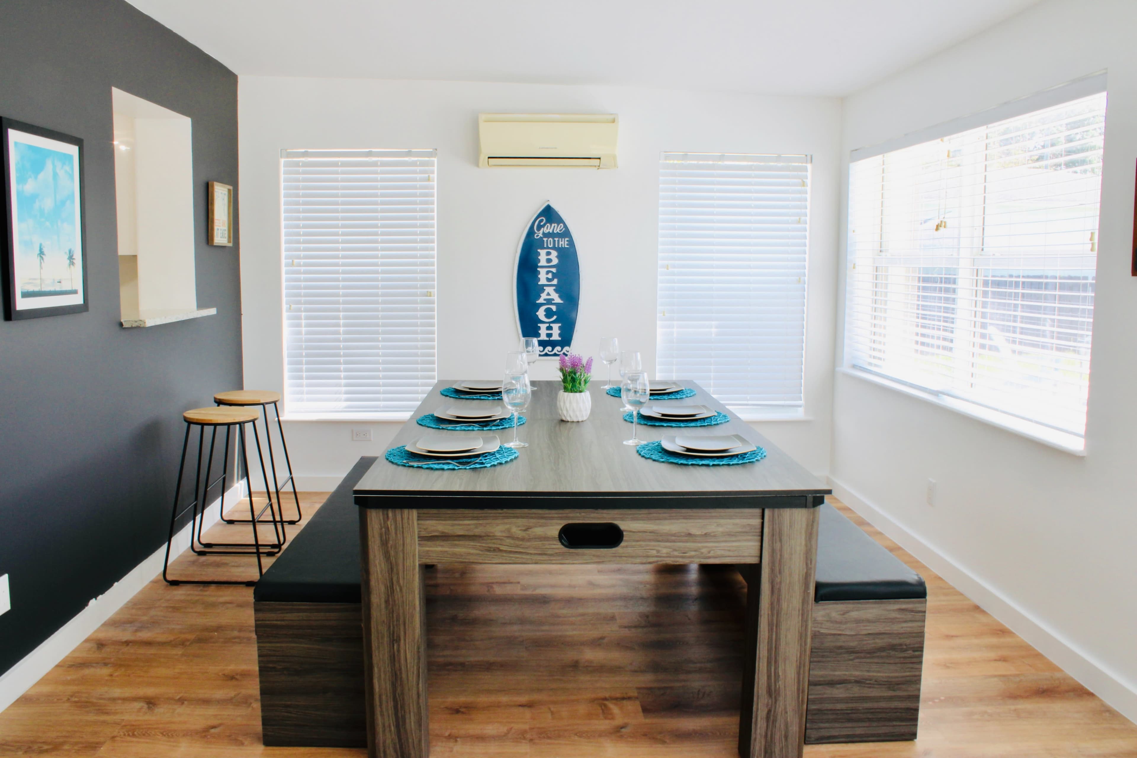 A dining area features a large wooden table with a surfboard-themed wall decoration and windows with blinds.