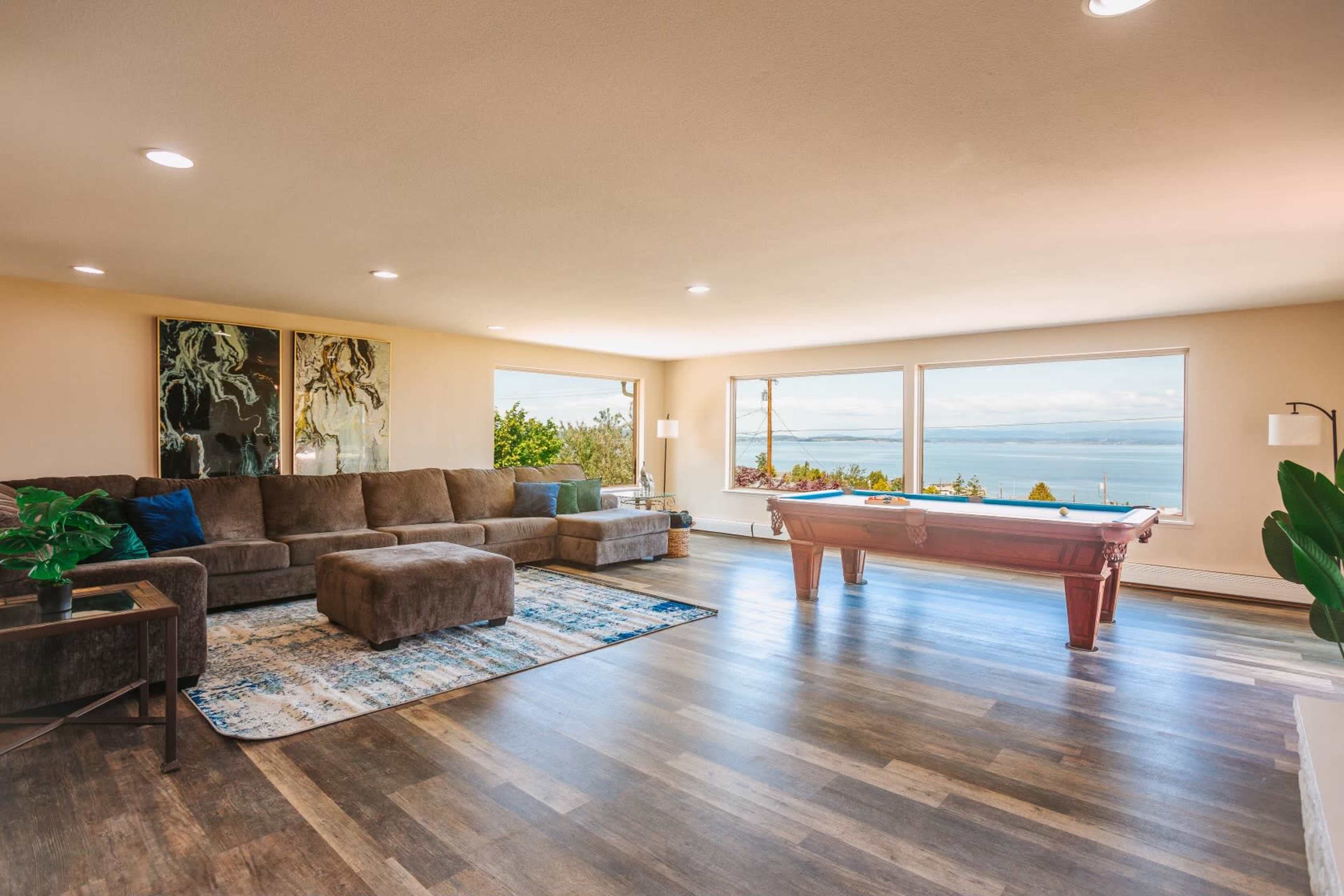 The spacious living area features a large sectional sofa, a billiard table, and expansive windows with a view of the water.