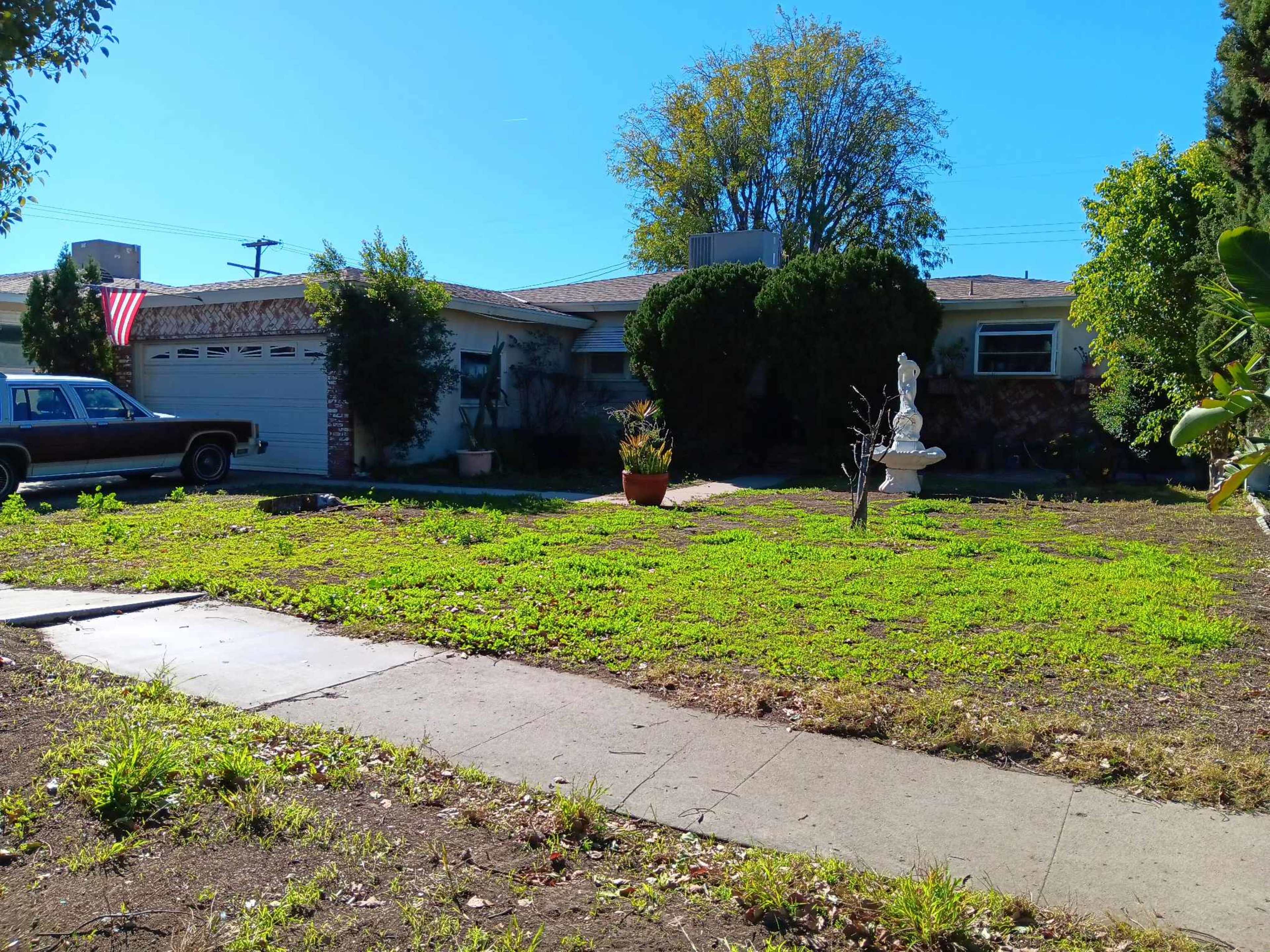 A grassy front yard features a statue and a potted plant in front of a single-story house with an American flag displayed.