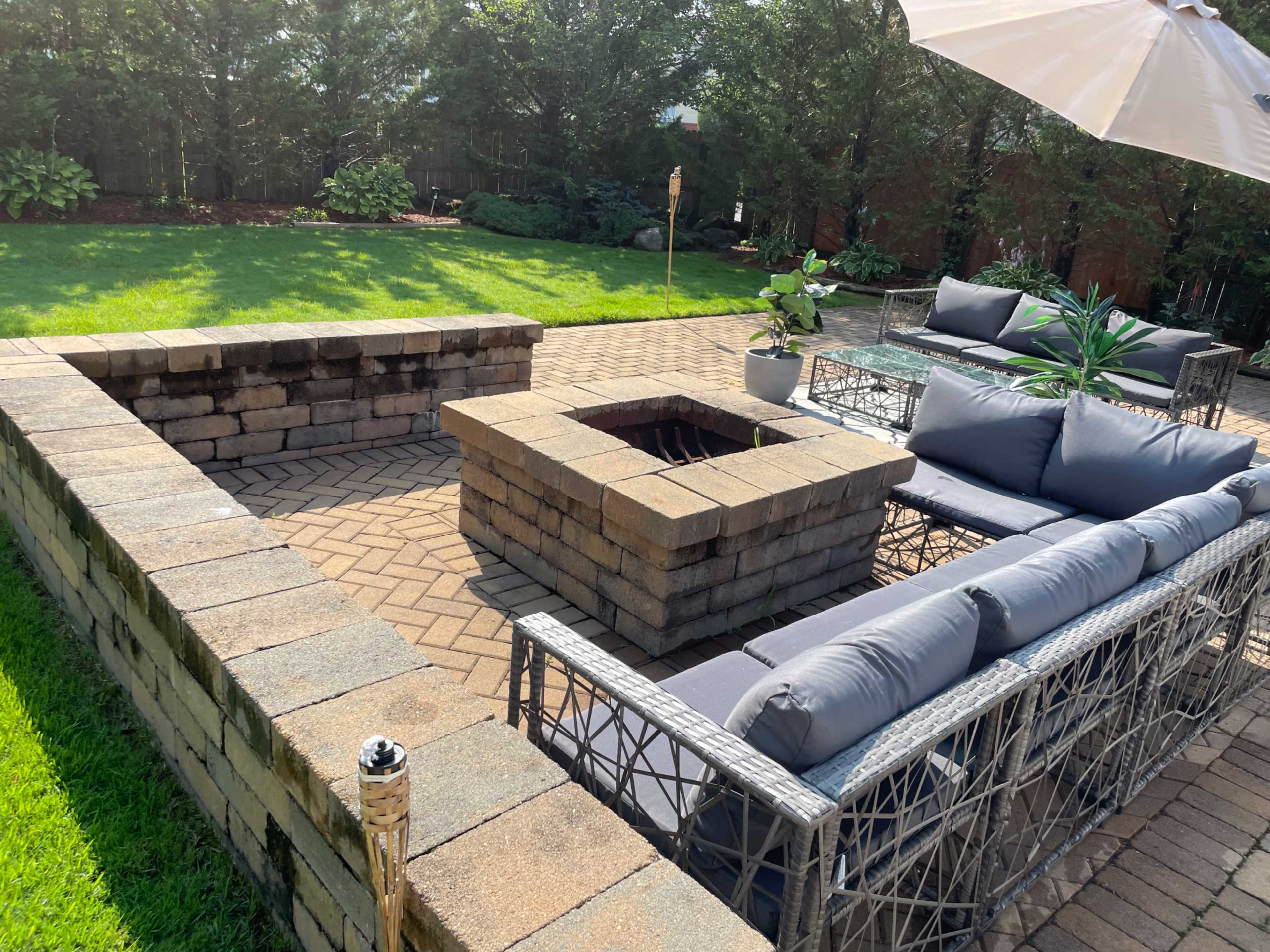 A patio area with a square fire pit surrounded by seating and a table, all situated on brick pavers.