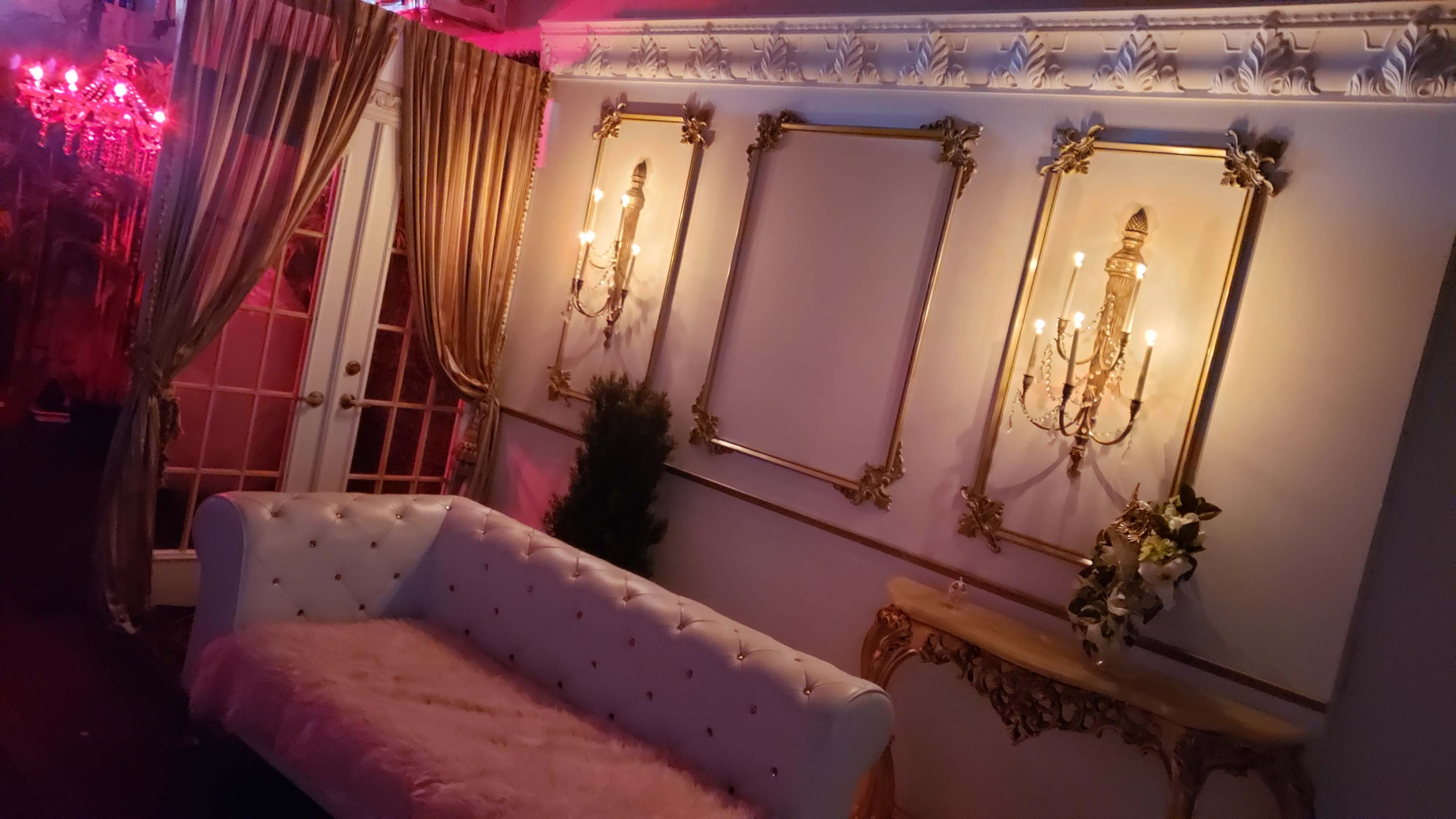 The scene features a luxurious room with a white tufted sofa, ornate wall sconce lighting, and decorative gold-framed empty picture spaces against a richly draped backdrop.