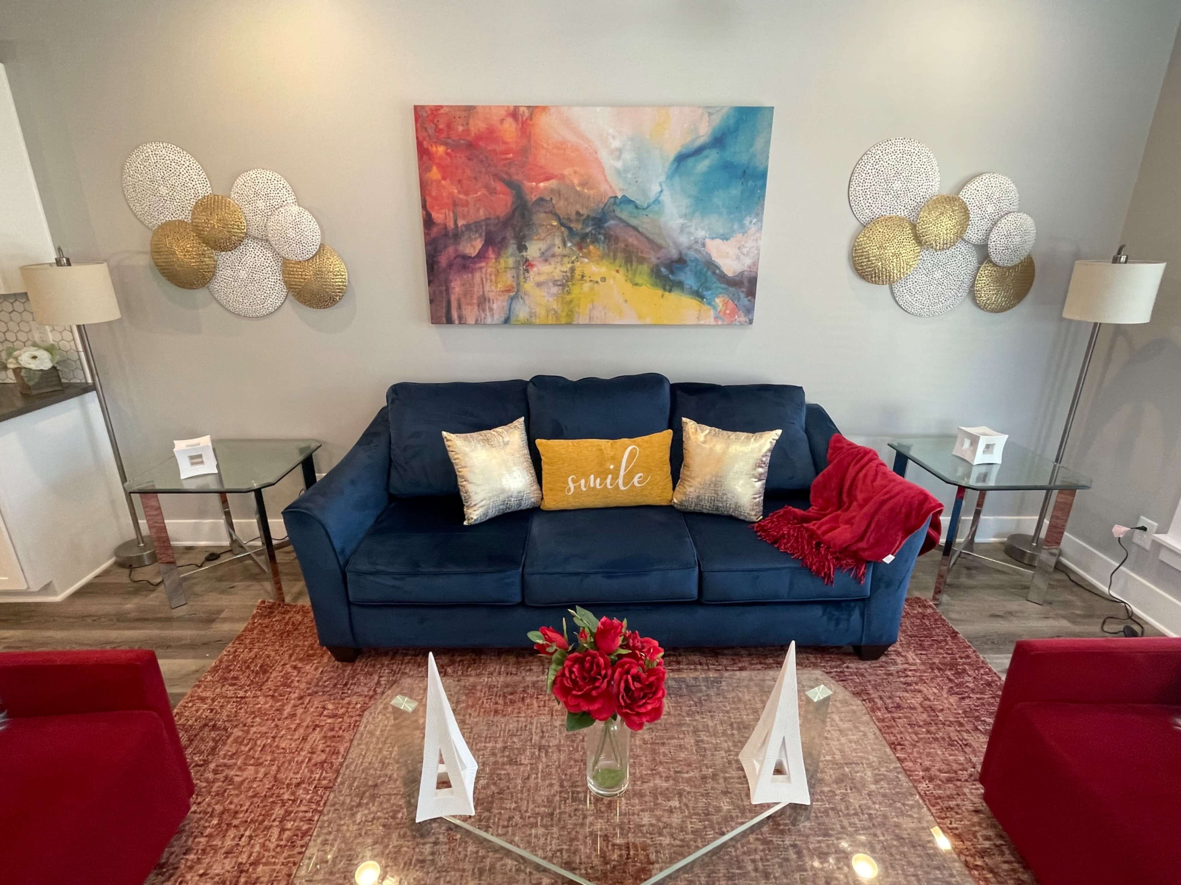 A blue sofa with decorative pillows and a red throw sits in a living room adorned with modern artwork and decorative wall hangings, flanked by glass tables and a floral centerpiece.