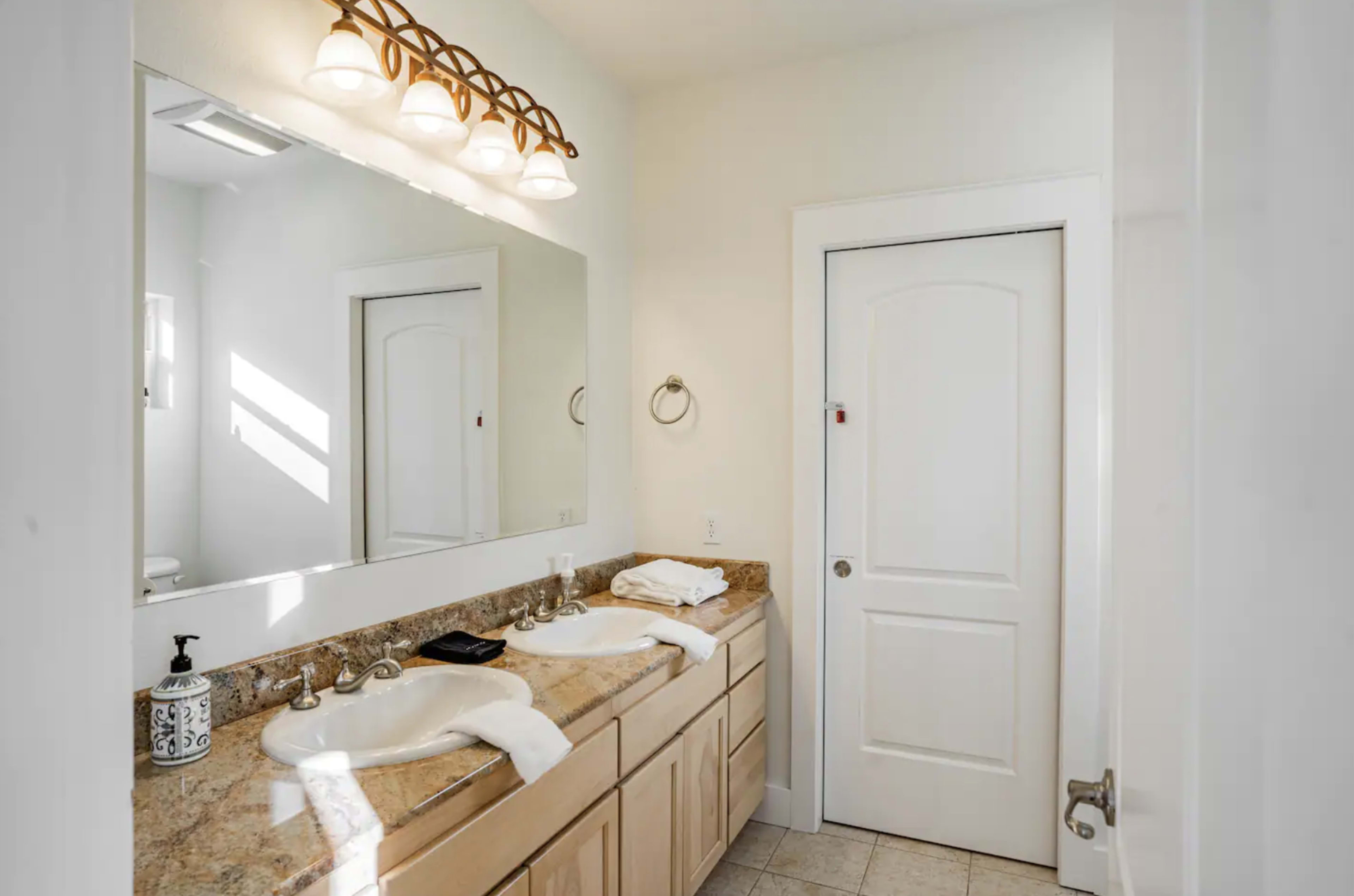 The image shows a bathroom with dual sinks, a large mirror, and a door leading to another area.