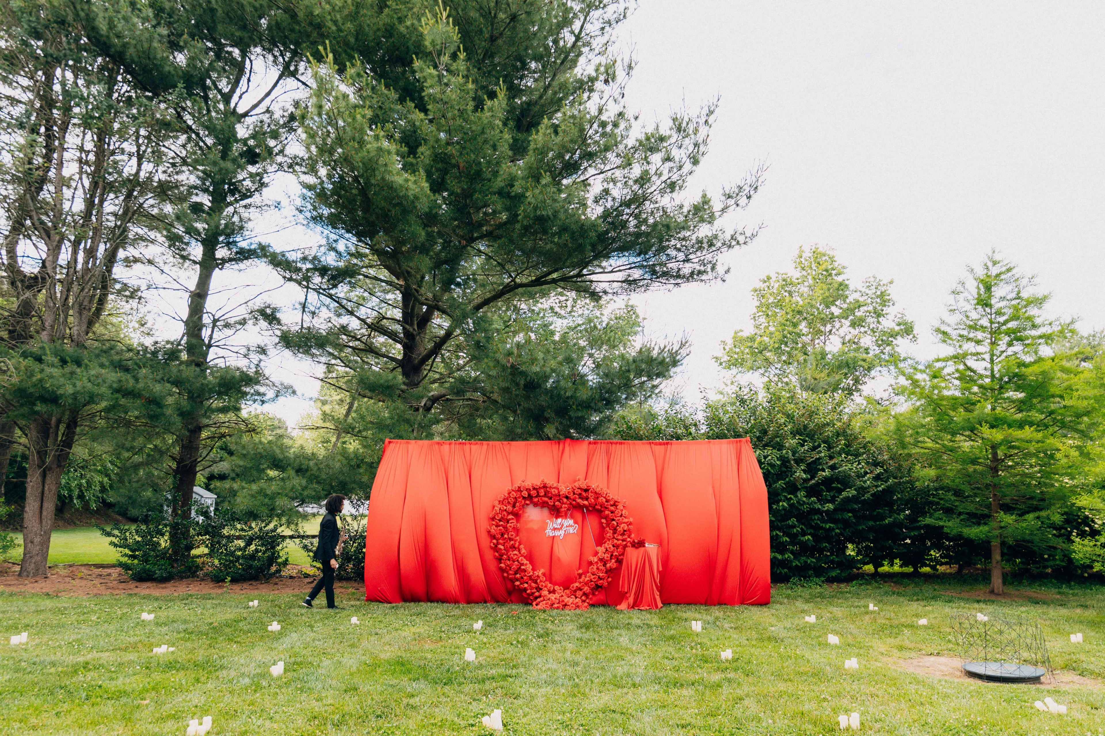 A person dressed in black walks past a large red backdrop shaped like a heart, situated in a grassy area surrounded by trees.