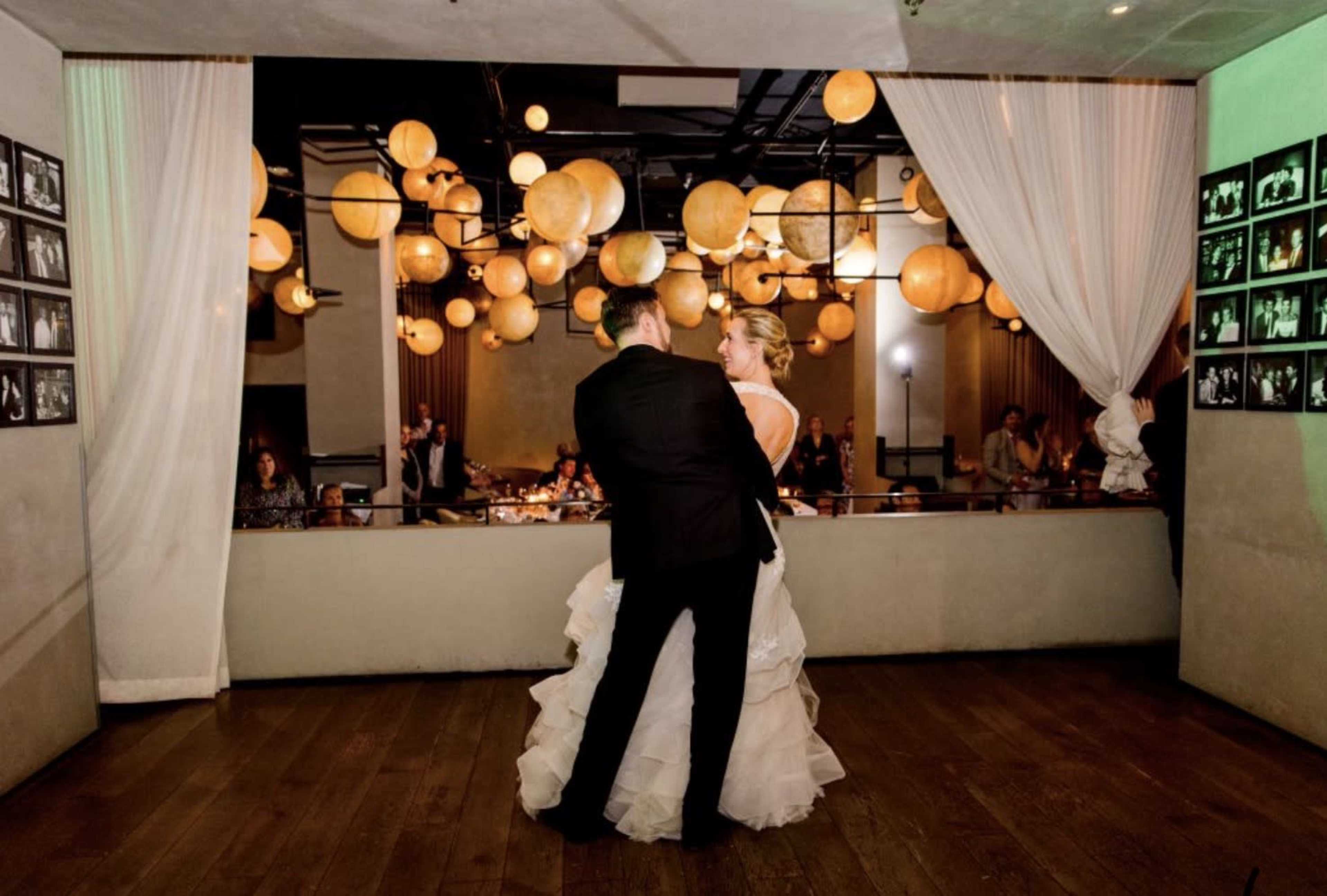 A couple dances on a wooden floor in a decorated venue filled with glowing round lanterns and framed photographs on the walls.