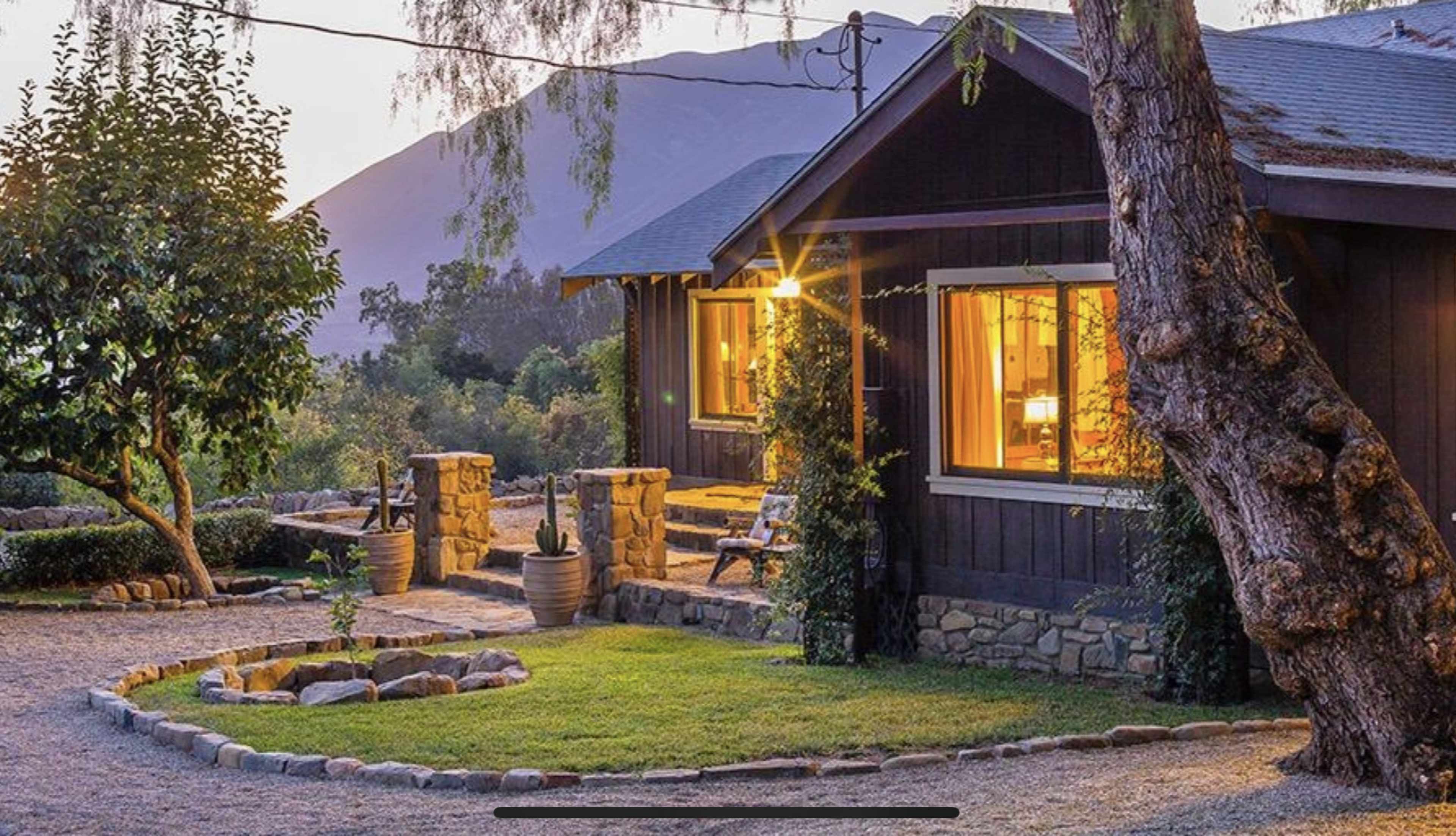 A quaint wooden cottage with warm lights visible through the windows is surrounded by a gravel pathway and a landscaped yard.