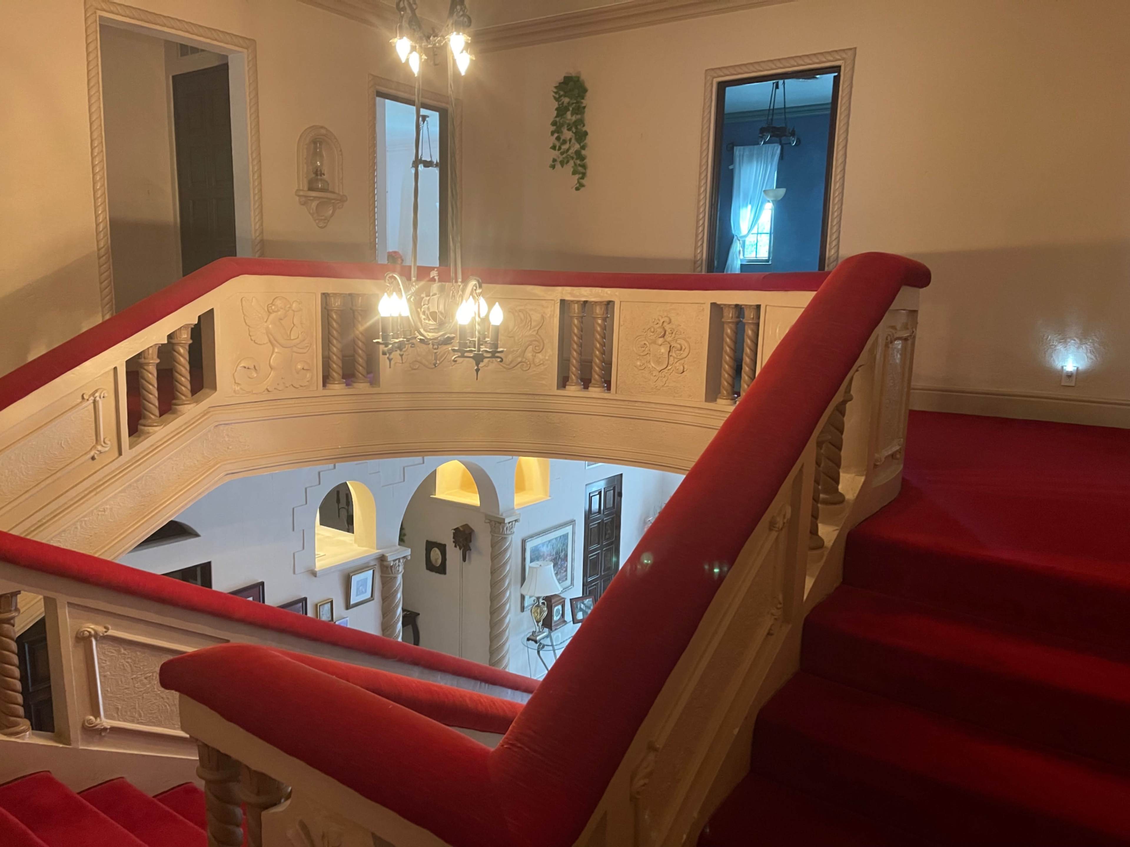 The image shows a grand staircase with red carpet and ornate railing, leading to an upper floor in a well-lit hallway.