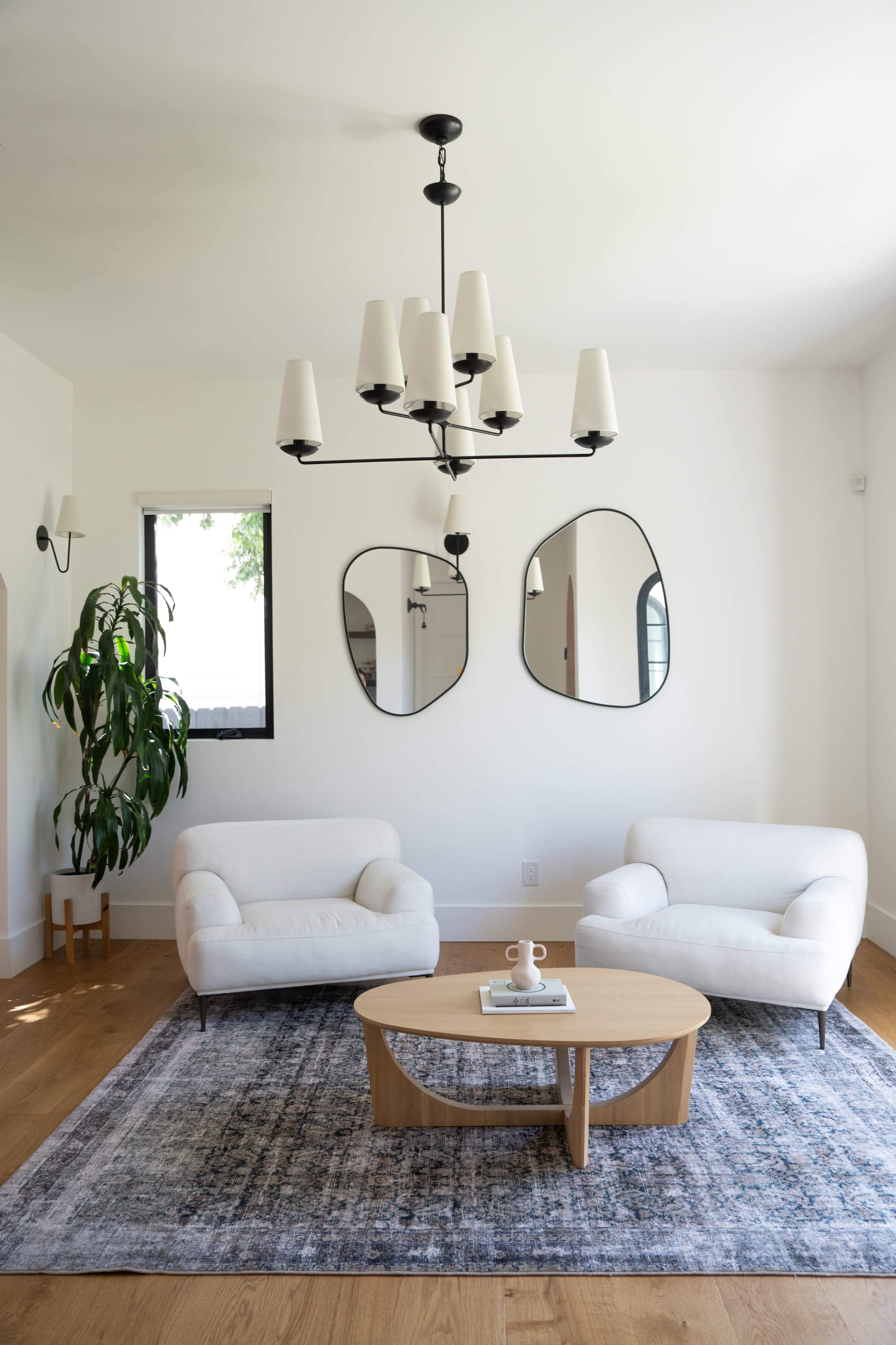 A modern living room features two white armchairs, a round wooden coffee table, a patterned rug, and a chandelier, complemented by two mirrors and a plant.