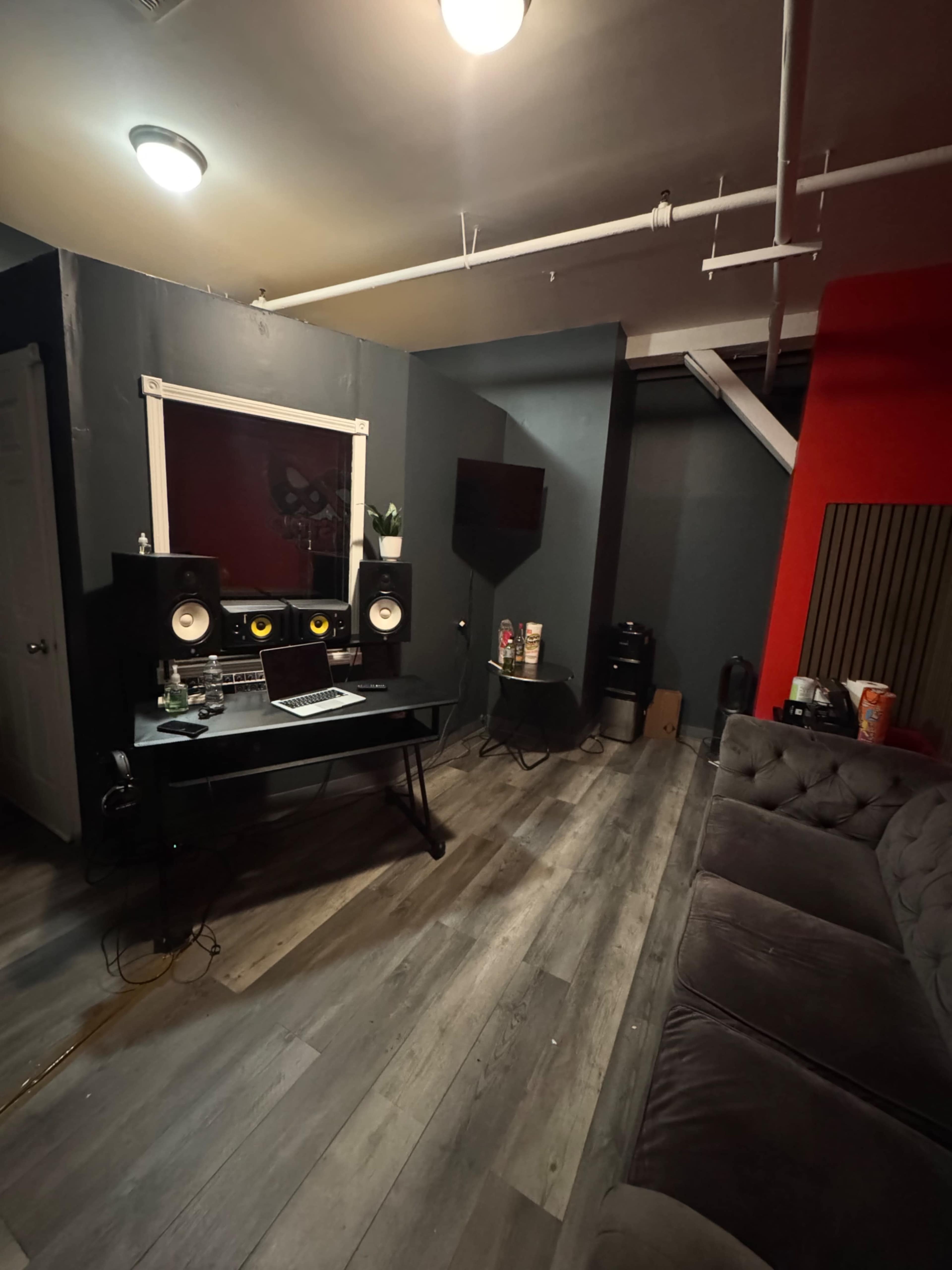 The image shows a music recording studio featuring a desk with a computer, speakers, and headphones, surrounded by gray walls and a red accent wall.