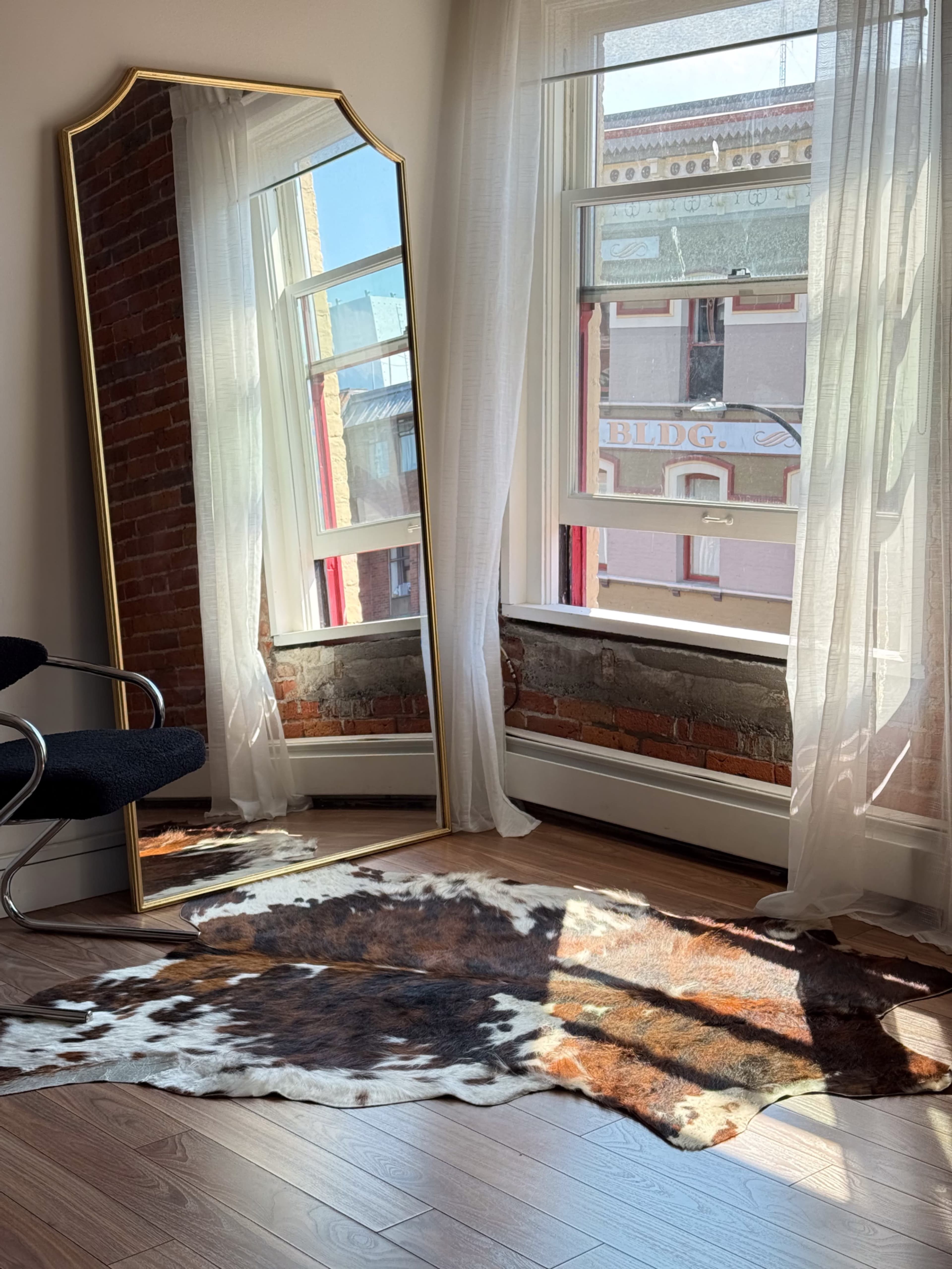 A large mirror stands next to a window with sheer curtains, casting a shadow on a patterned cowhide rug on the wooden floor.