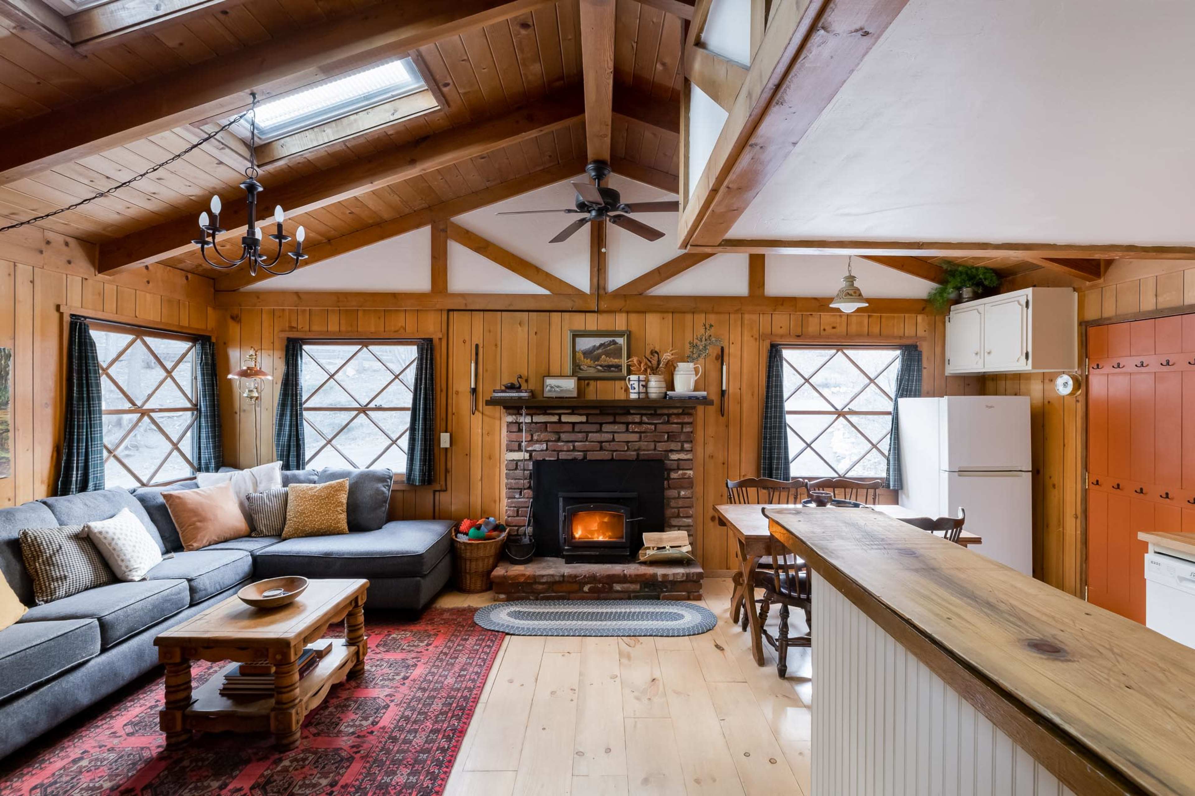 A cozy cabin interior featuring a living area with a brick fireplace, a sectional sofa, and a dining table near a kitchen with rustic decor.