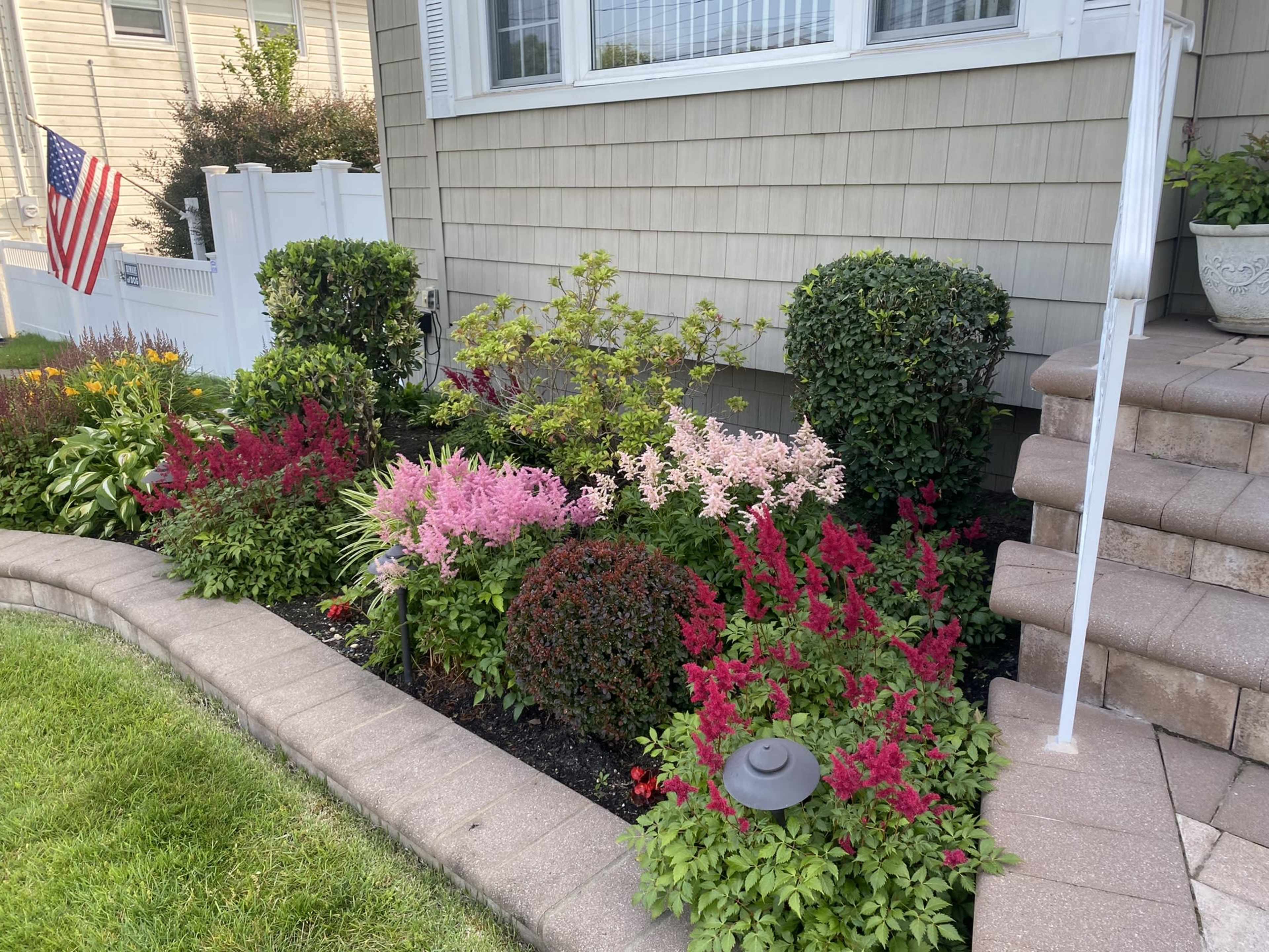 A well-maintained garden featuring a variety of colorful flowers and shrubs along a stone pathway leading to a house, with an American flag visible in the background.
