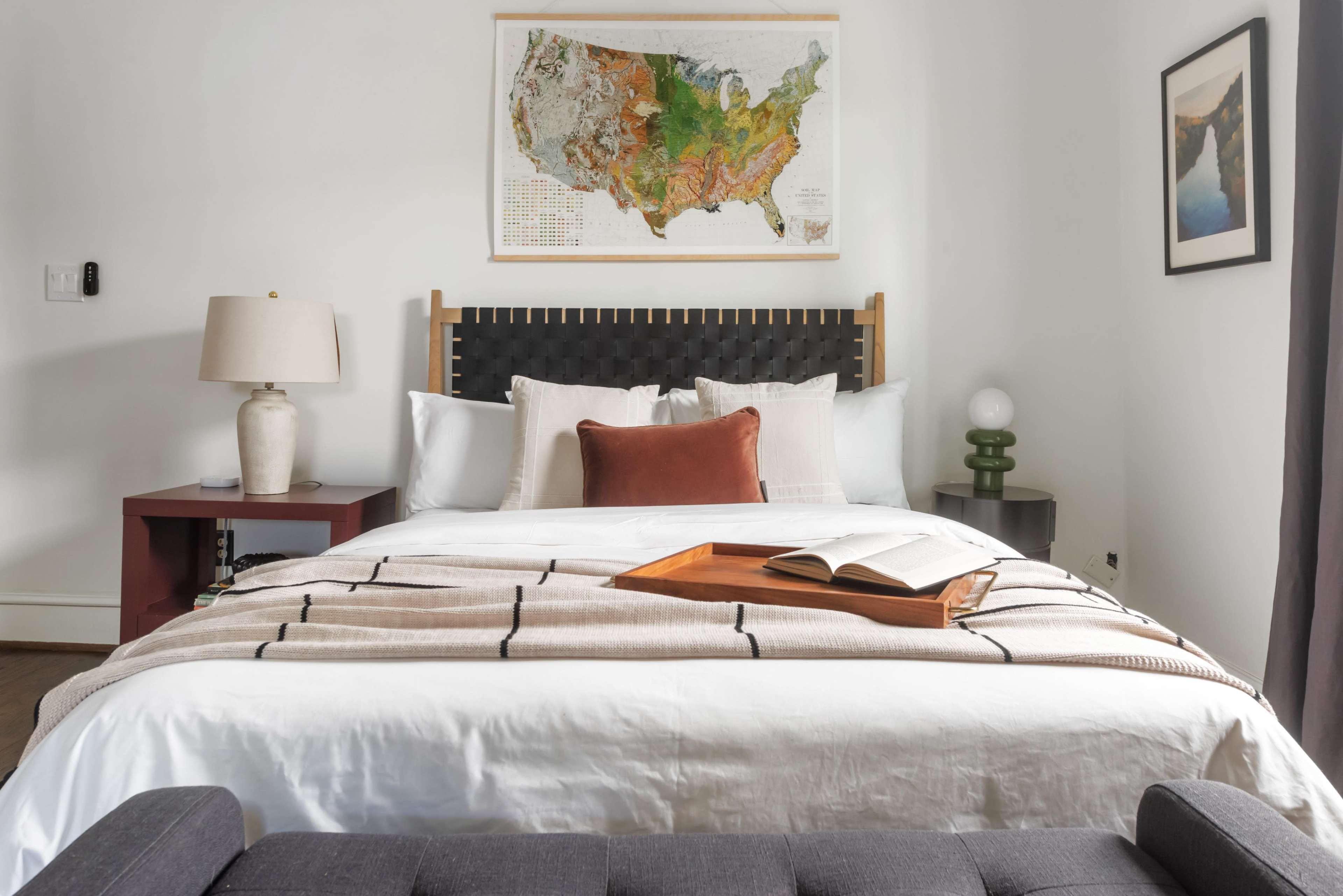 The scene shows a neatly arranged bedroom with a bed featuring a white comforter, a decorative throw, and a wall-mounted map of the United States above the headboard.