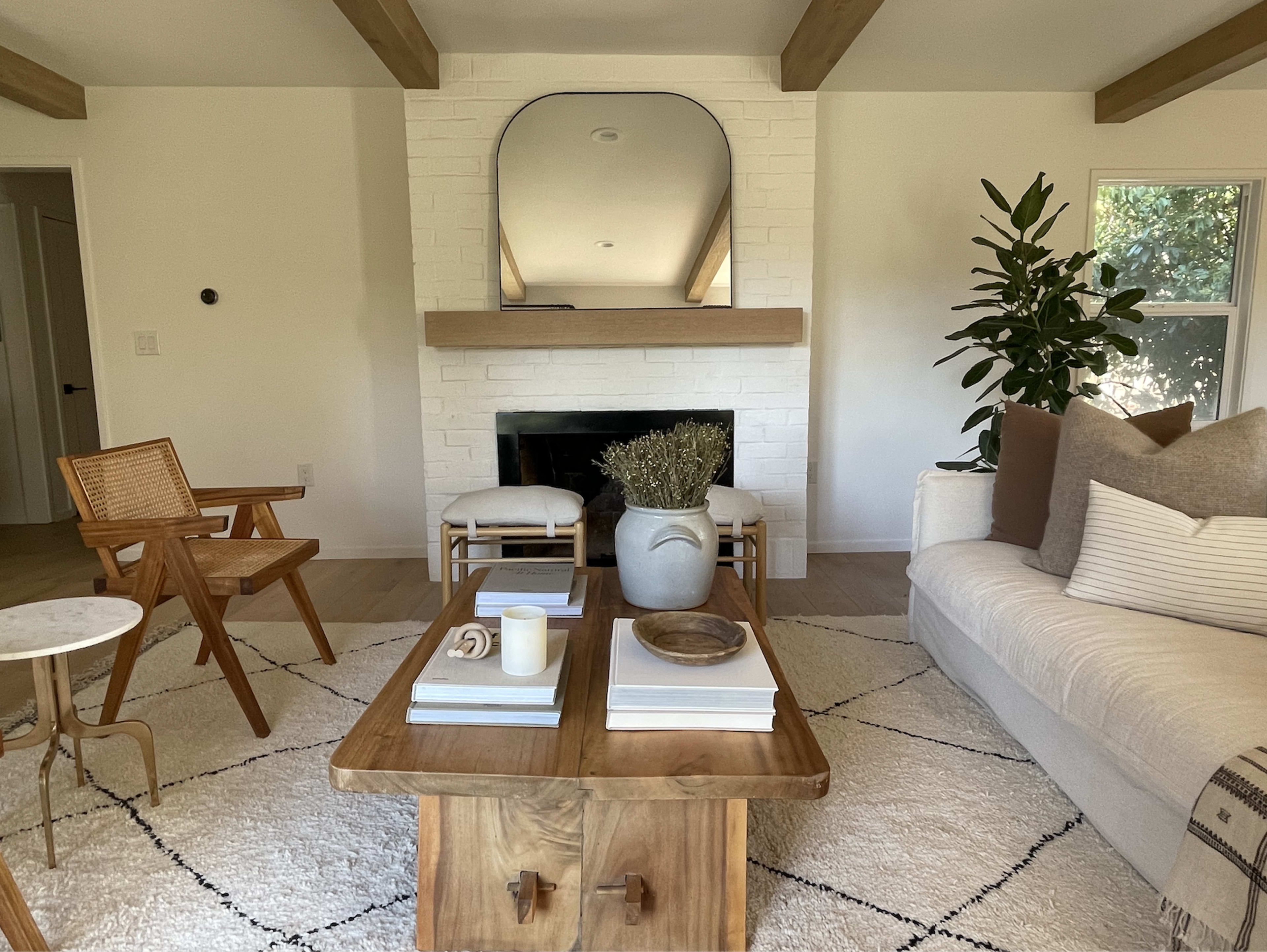 The image shows a living room featuring a white sofa, a wooden coffee table, and a minimalist fireplace with a large mirror above it surrounded by plants.