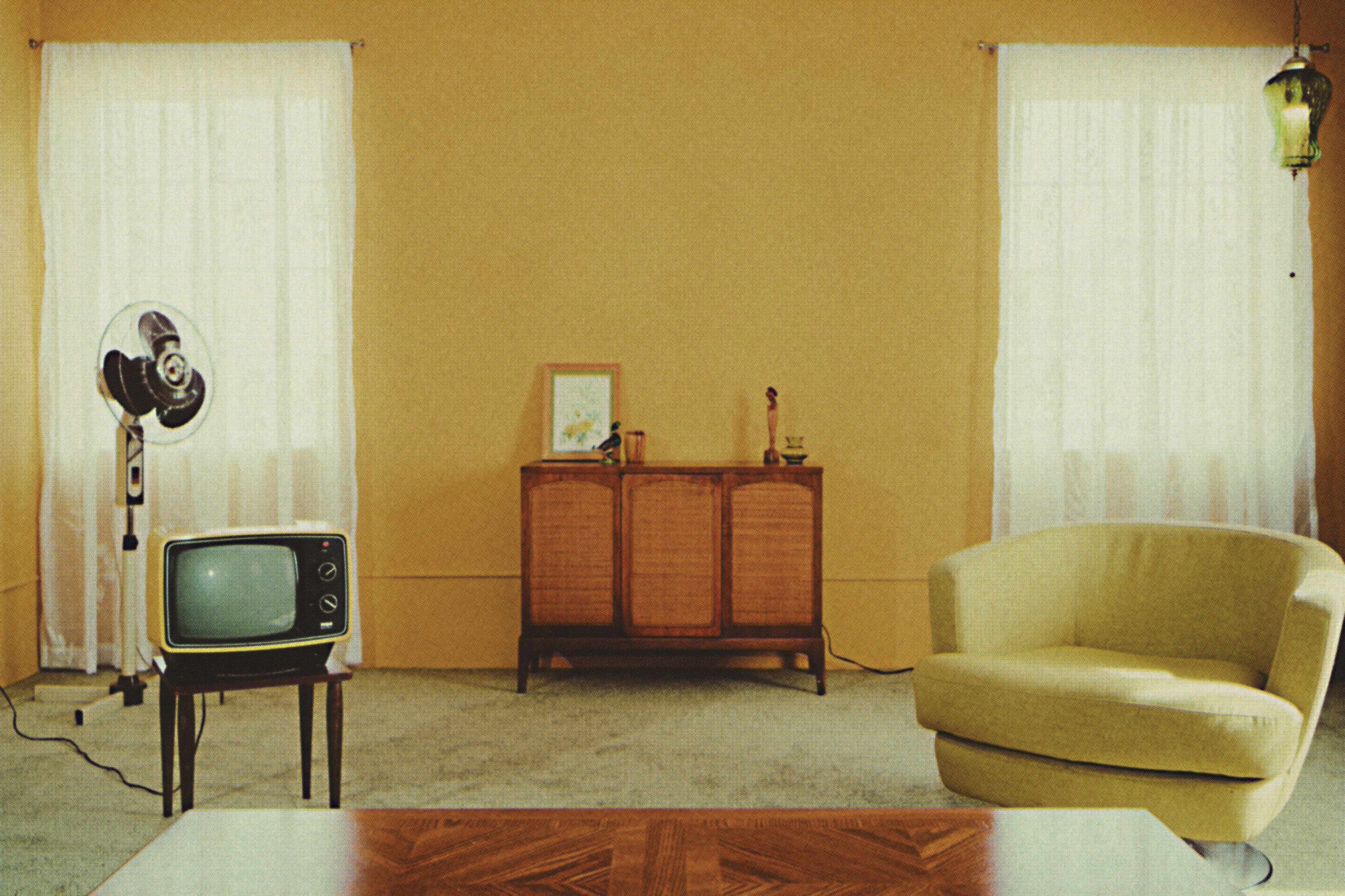 A sparsely furnished room featuring a vintage television, a retro fan, a wooden cabinet, and a yellow armchair, all set against light-colored walls and large windows.