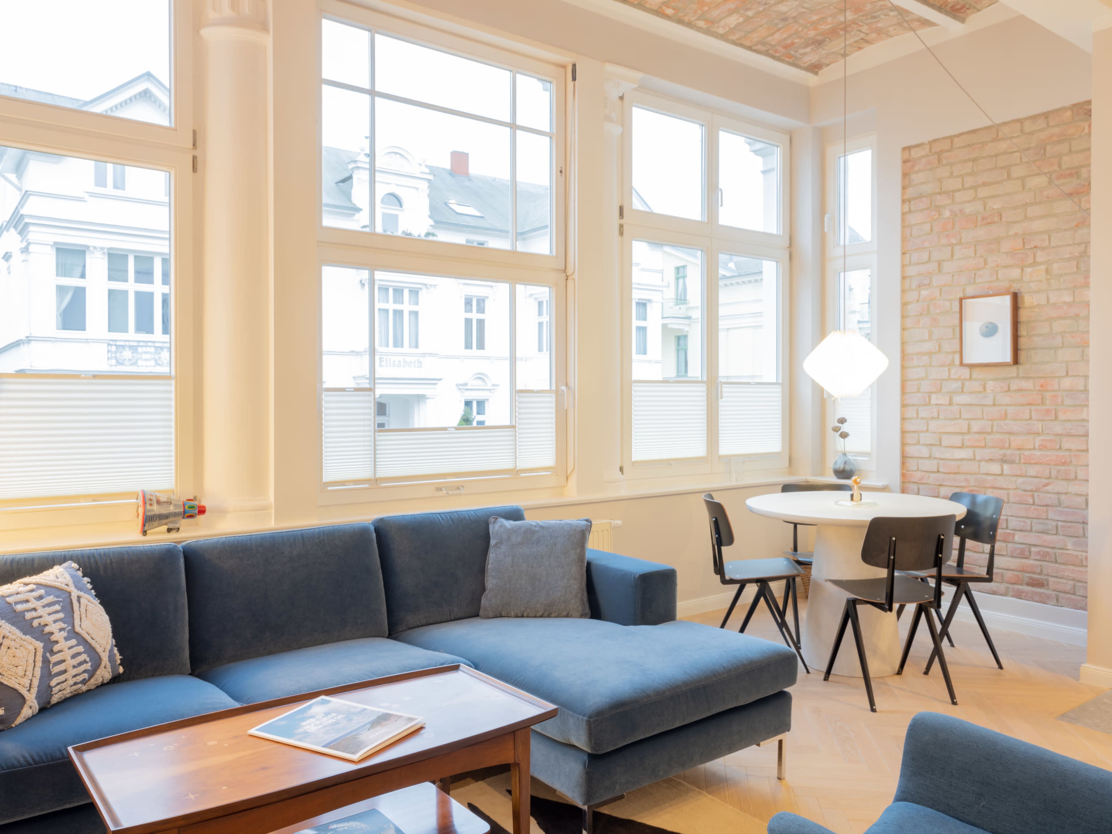 A well-lit living room features a blue sectional couch, a wooden coffee table, and a round dining table surrounded by black chairs, with large windows showing a view of the outside.