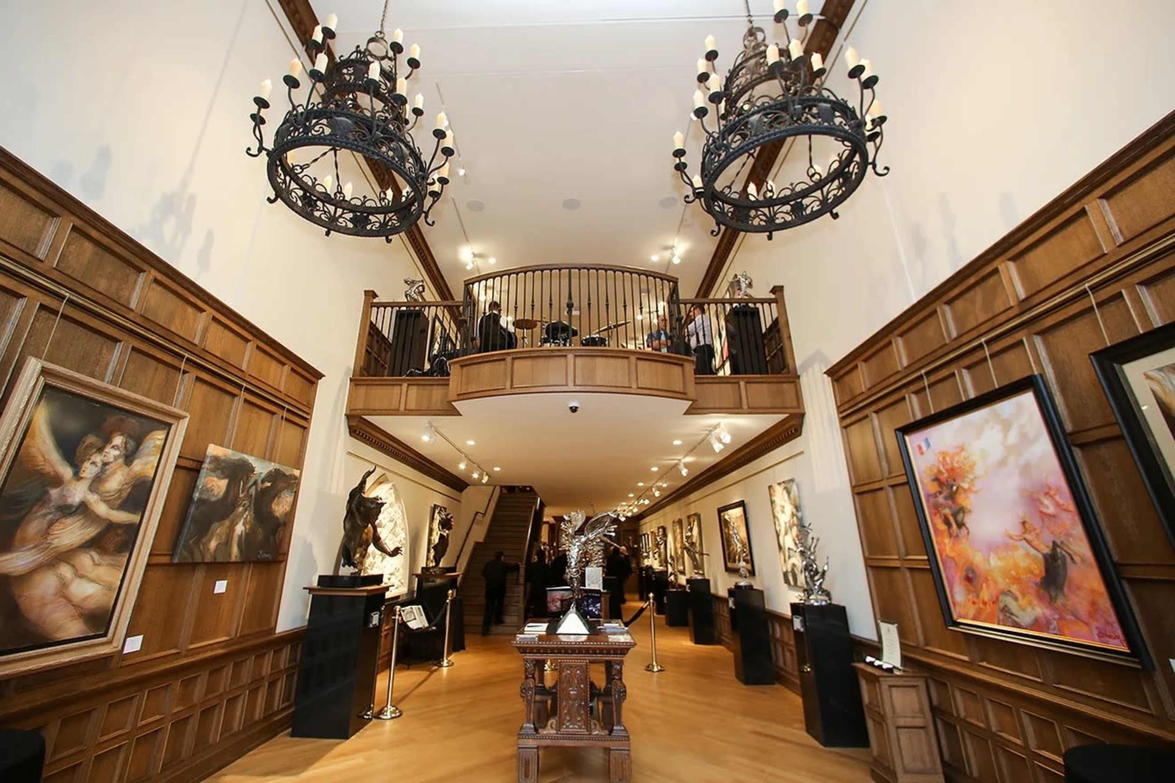 The interior of an art gallery features wooden paneling, multiple large paintings, and a staircase leading to a second level.