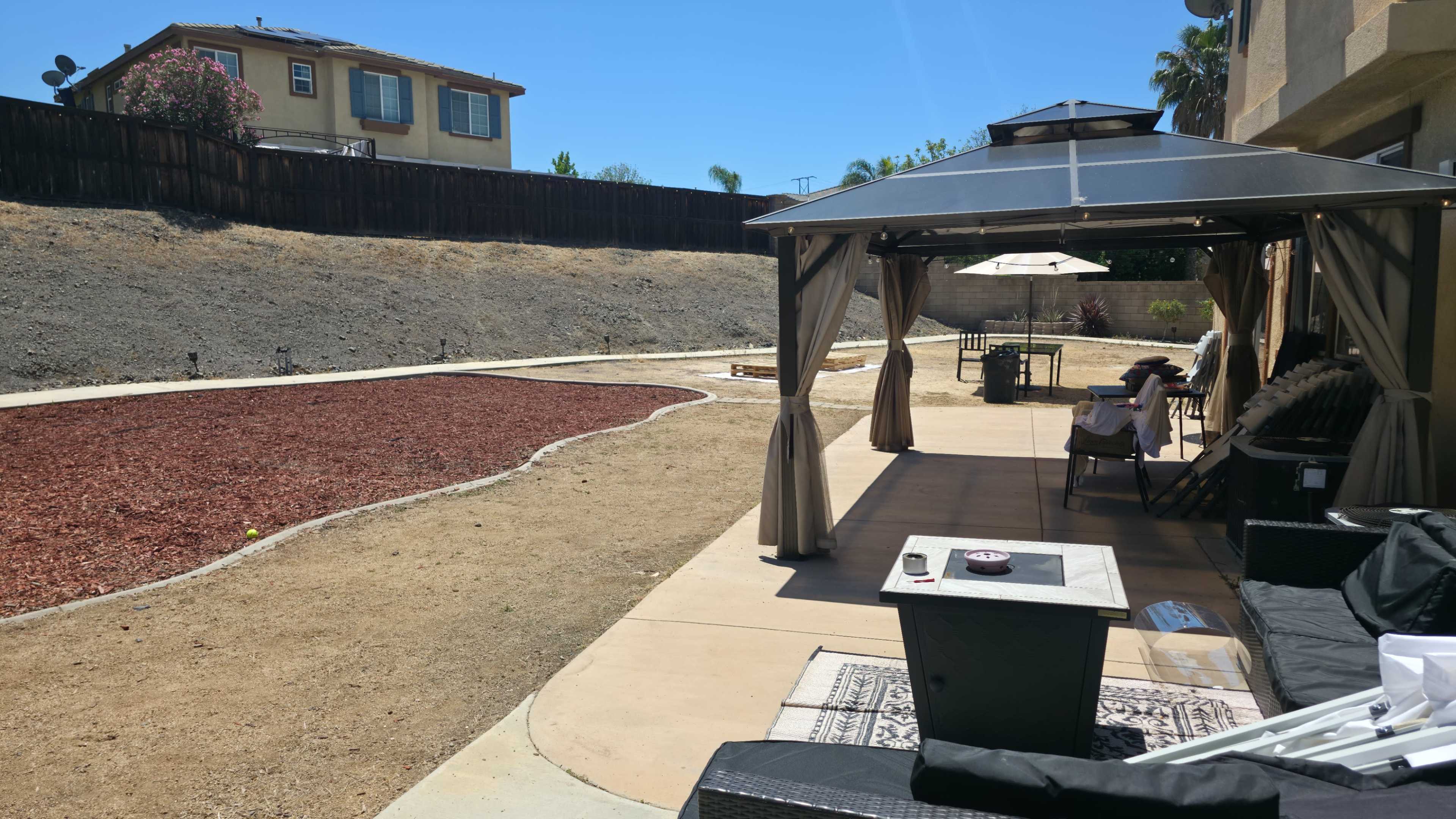 The image shows a backyard with a gravel area, a gazebo, and a patio set against a backdrop of a barren hill and a residential building.
