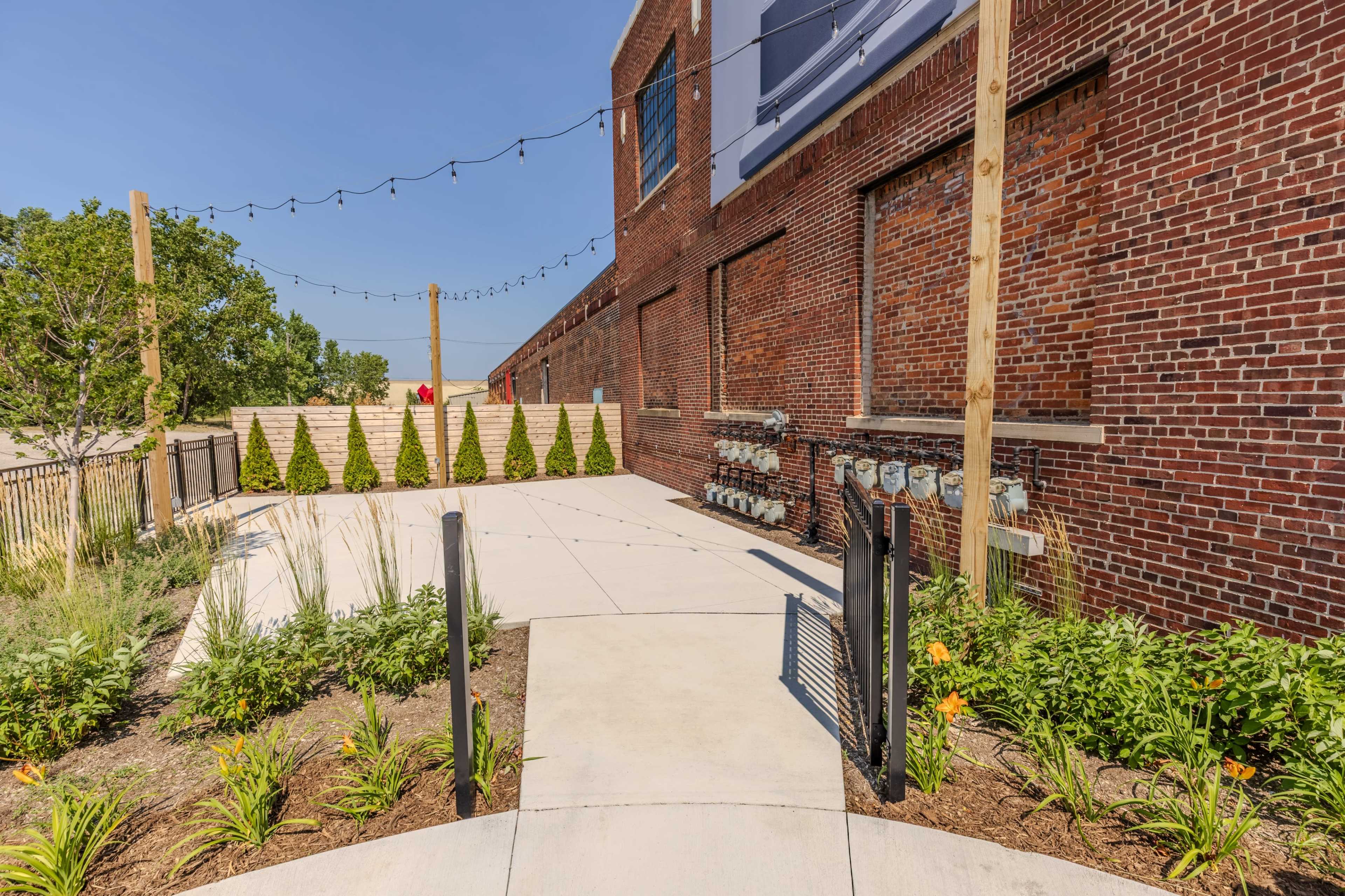 The image shows a paved pathway leading to a brick building, flanked by neatly trimmed shrubs and outdoor lighting.