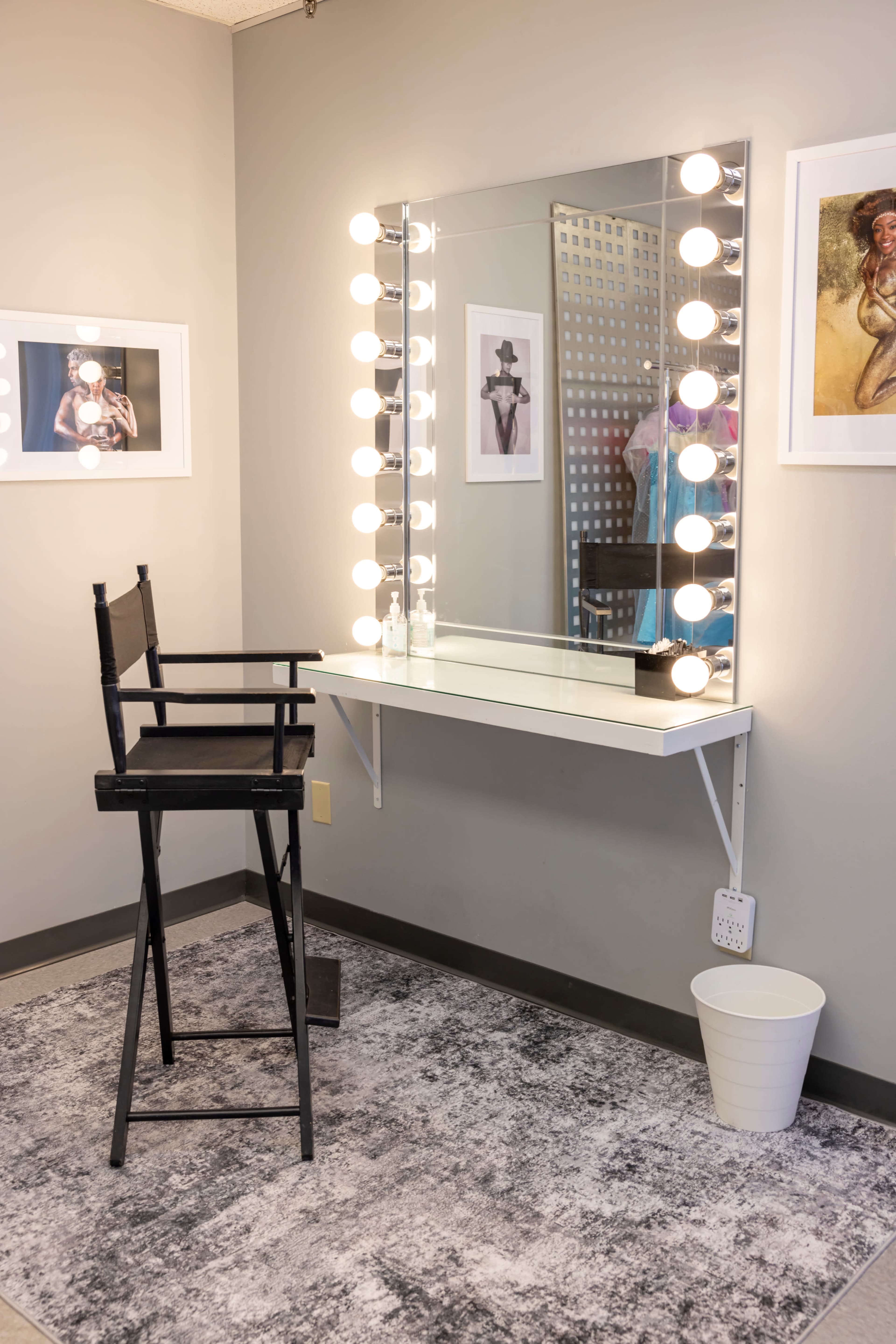 The image shows a makeup vanity area with a large mirror framed by bright bulbs, a high stool, and framed photos on the wall.